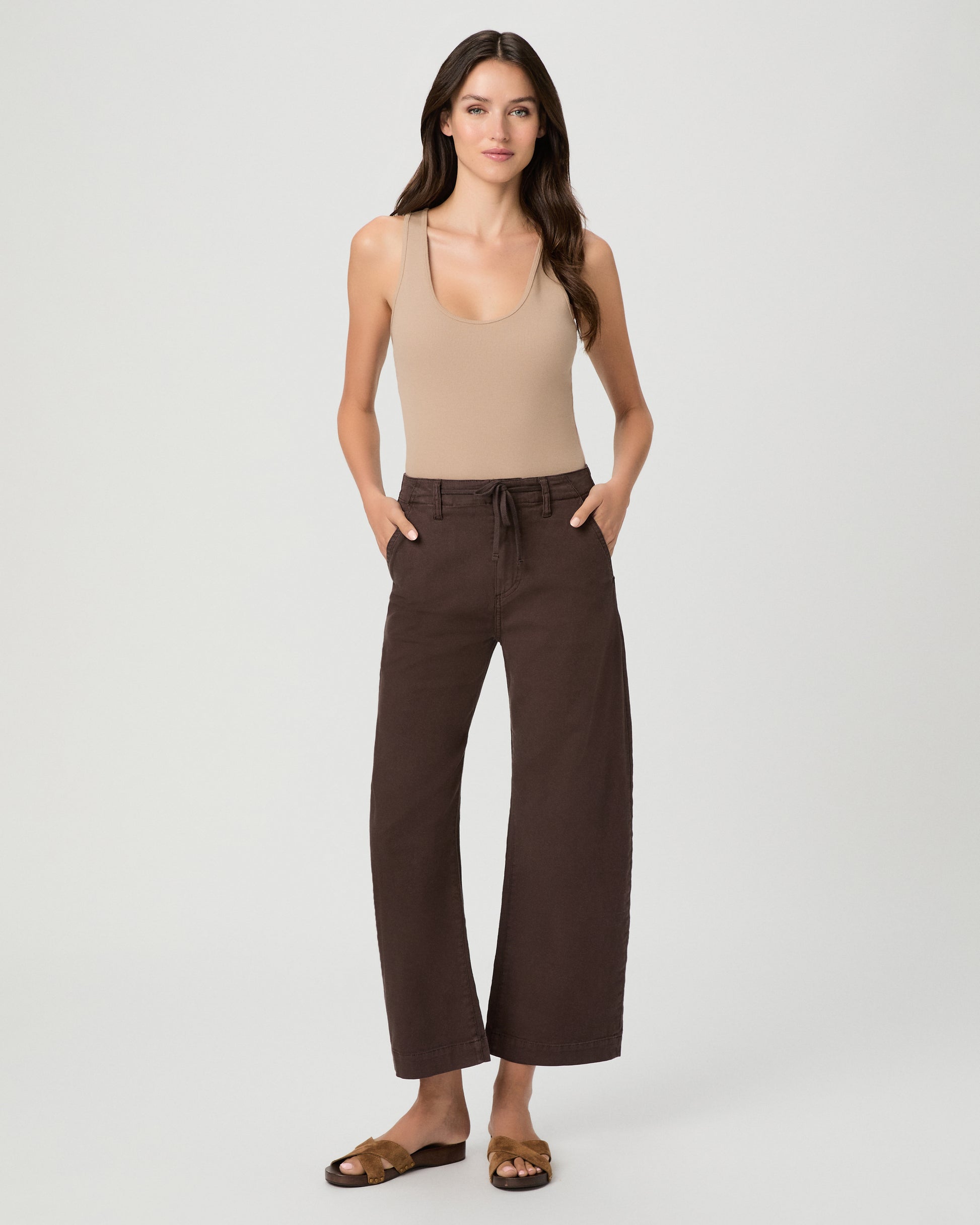 A woman modeling a pair of dark brown wide-leg cropped pants with a drawstring waist, paired with a beige tank top and brown sandals, against a light gray studio background.