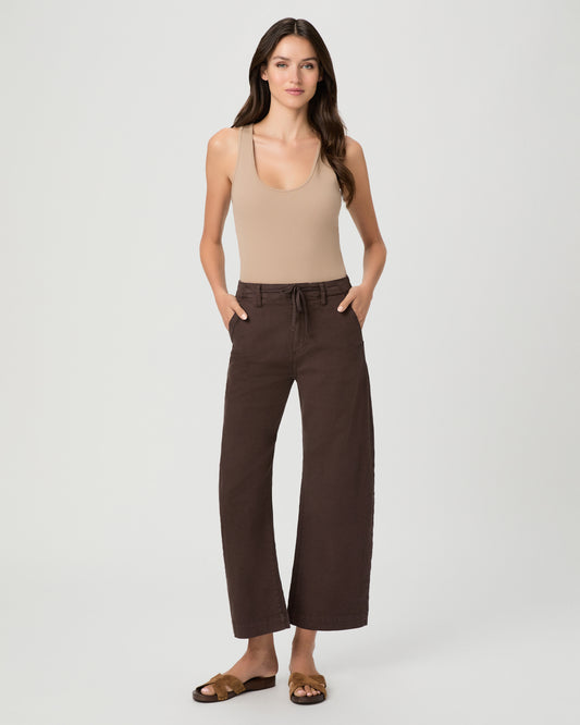 A woman modeling a pair of dark brown wide-leg cropped pants with a drawstring waist, paired with a beige tank top and brown sandals, against a light gray studio background.