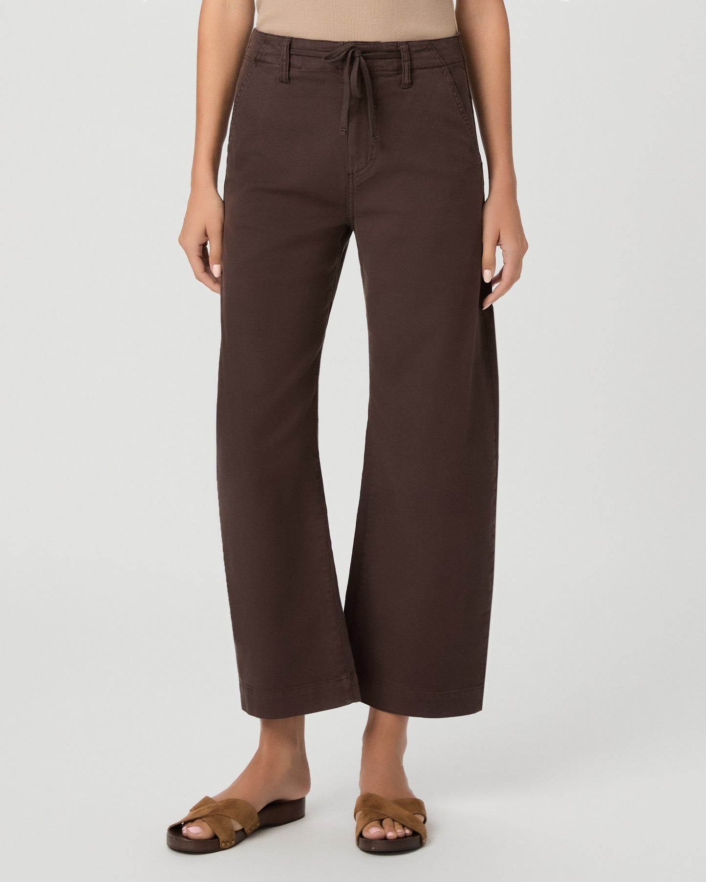 A woman modeling a pair of dark brown wide-leg cropped pants with a drawstring waist, paired with a beige tank top and brown sandals, against a light gray studio background.