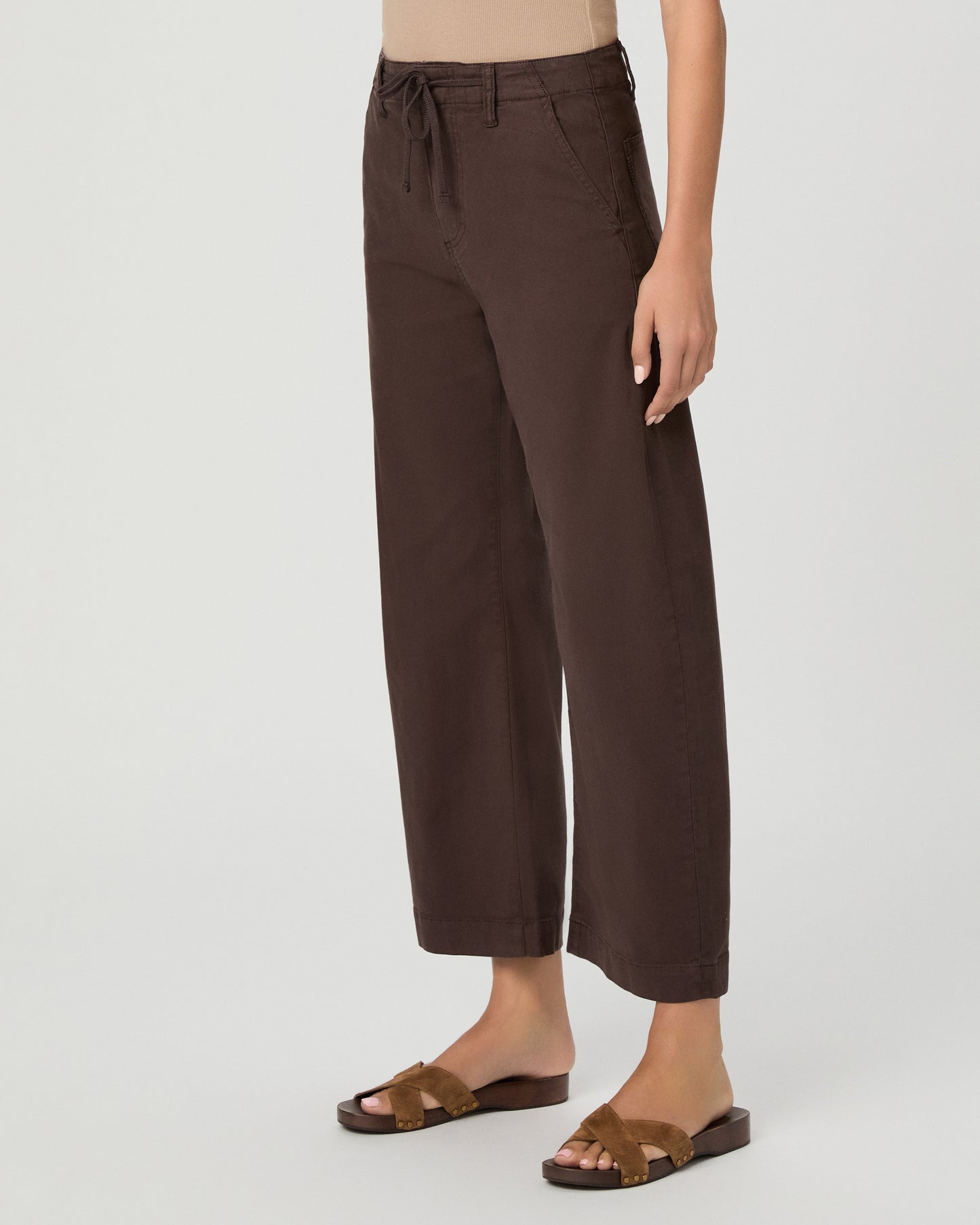 A woman modeling a pair of dark brown wide-leg cropped pants with a drawstring waist, paired with a beige tank top and brown sandals, against a light gray studio background.
