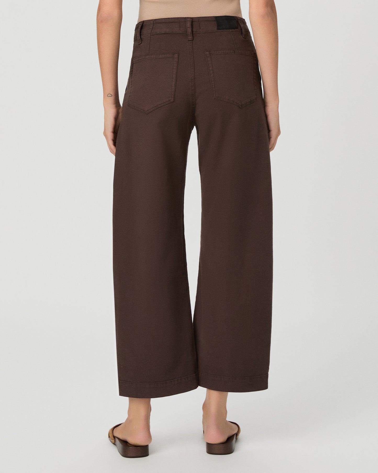 A woman modeling a pair of dark brown wide-leg cropped pants with a drawstring waist, paired with a beige tank top and brown sandals, against a light gray studio background.
