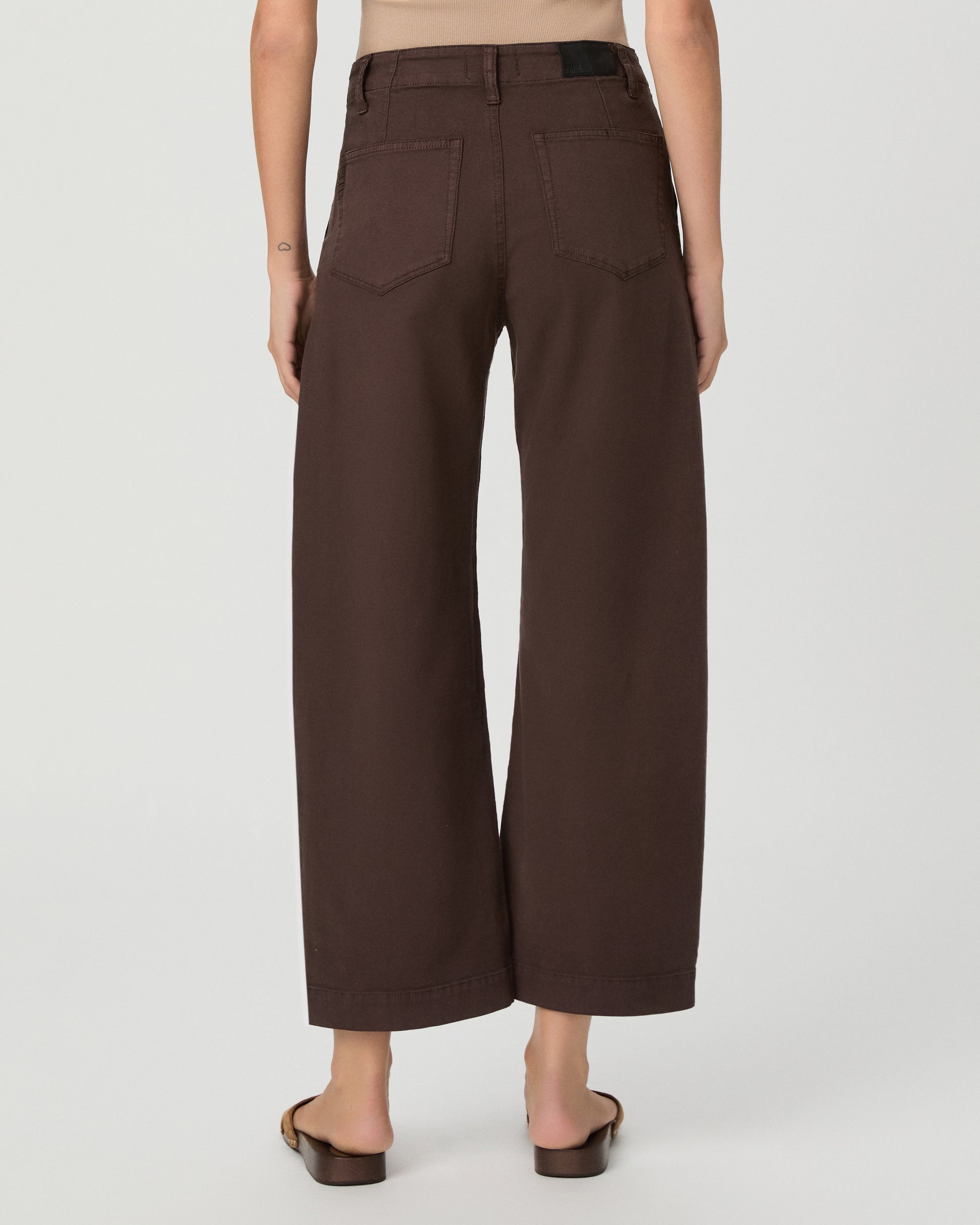 A woman modeling a pair of dark brown wide-leg cropped pants with a drawstring waist, paired with a beige tank top and brown sandals, against a light gray studio background.