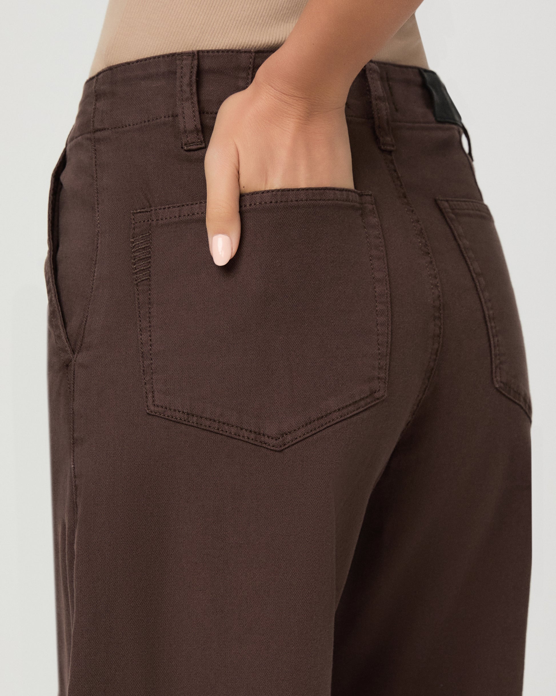 A woman modeling a pair of dark brown wide-leg cropped pants with a drawstring waist, paired with a beige tank top and brown sandals, against a light gray studio background.