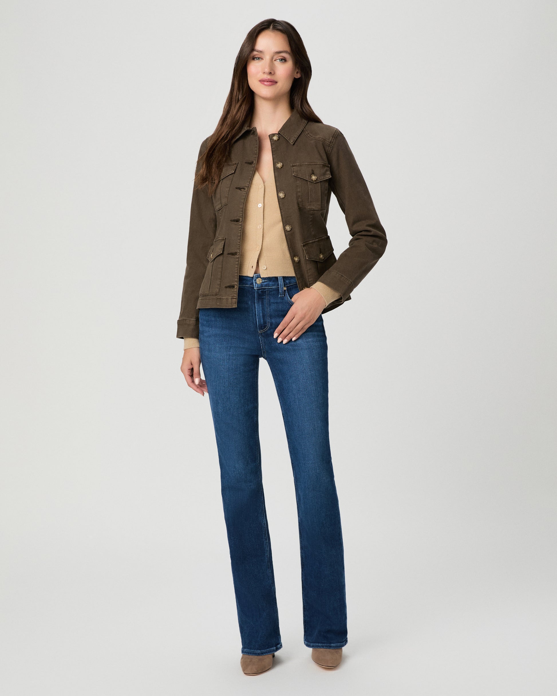 A woman modeling a dark brown utility jacket with four front flap pockets and gold-tone buttons, paired with blue jeans, against a light gray studio background.