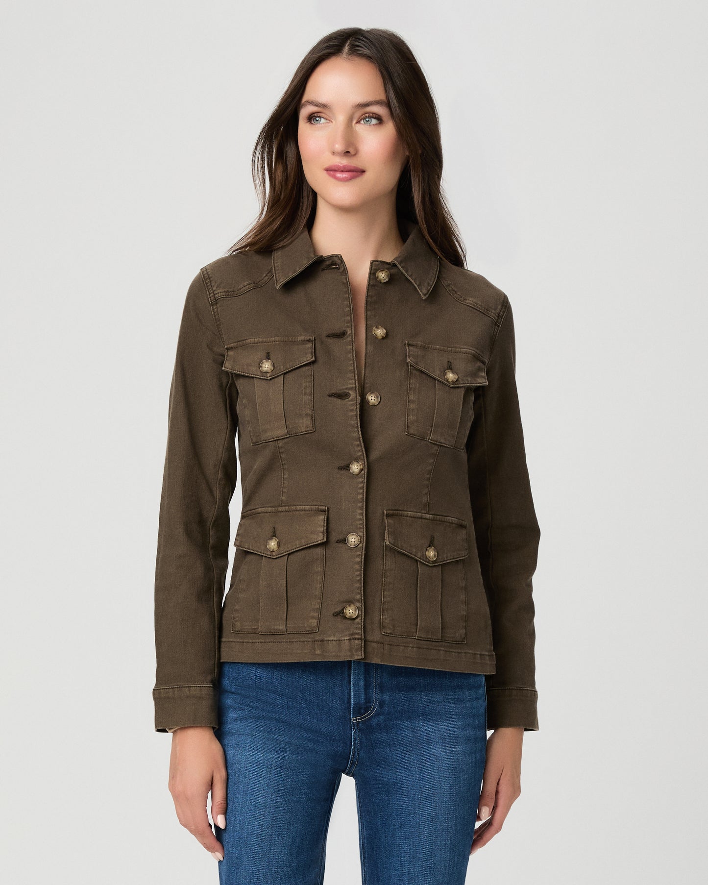 A woman modeling a dark brown utility jacket with four front flap pockets and gold-tone buttons, paired with blue jeans, against a light gray studio background.