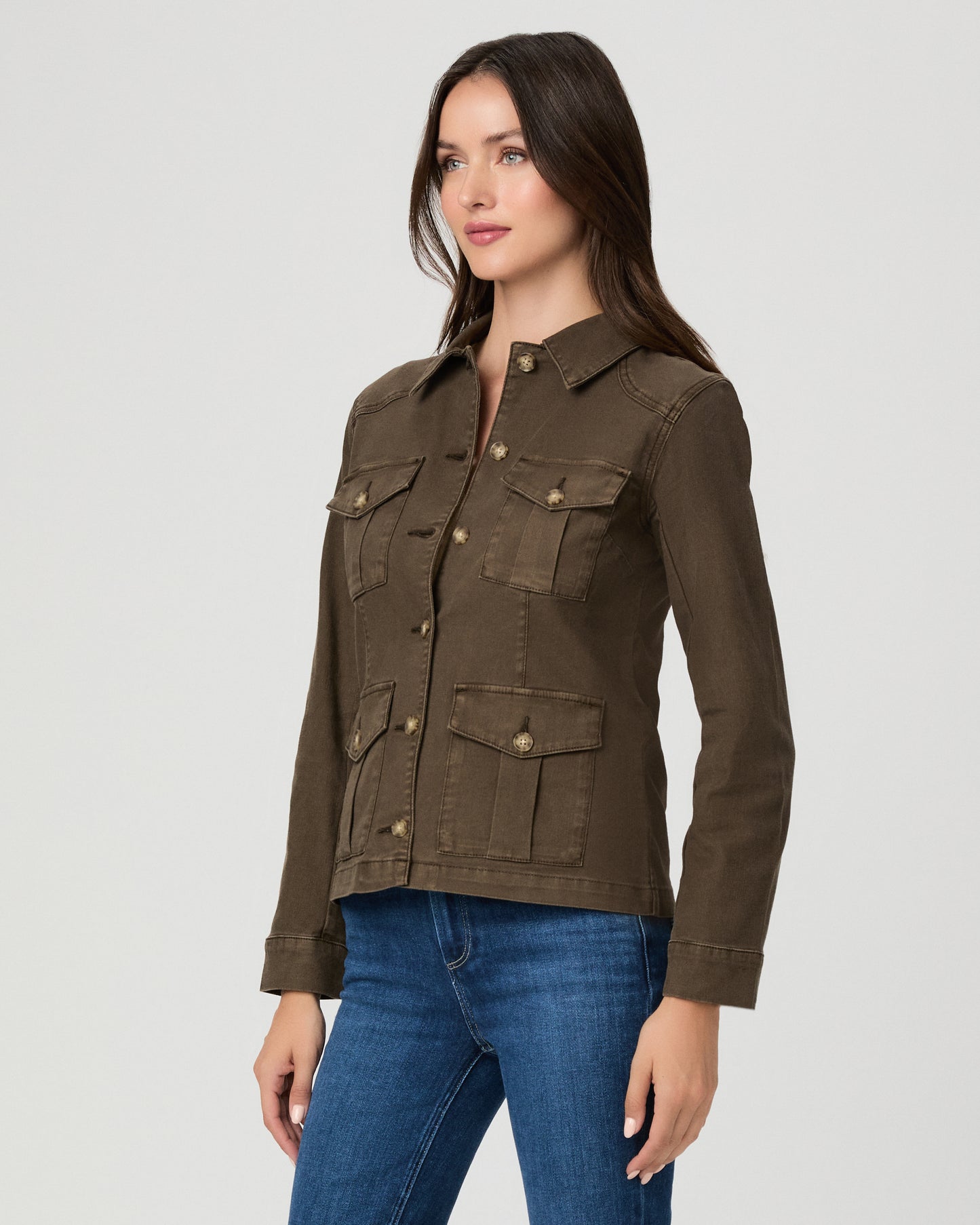 A woman modeling a dark brown utility jacket with four front flap pockets and gold-tone buttons, paired with blue jeans, against a light gray studio background.