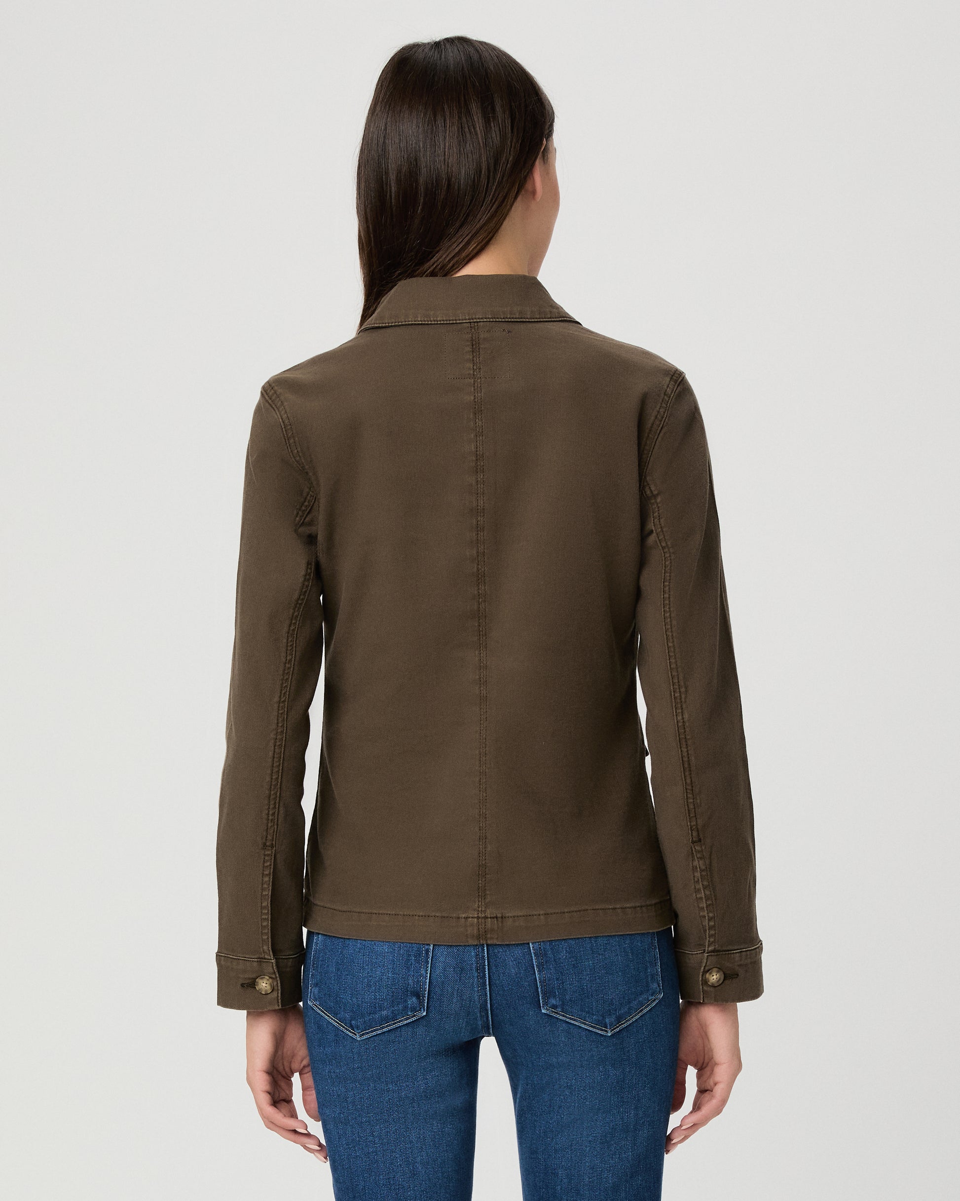 A woman modeling a dark brown utility jacket with four front flap pockets and gold-tone buttons, paired with blue jeans, against a light gray studio background.