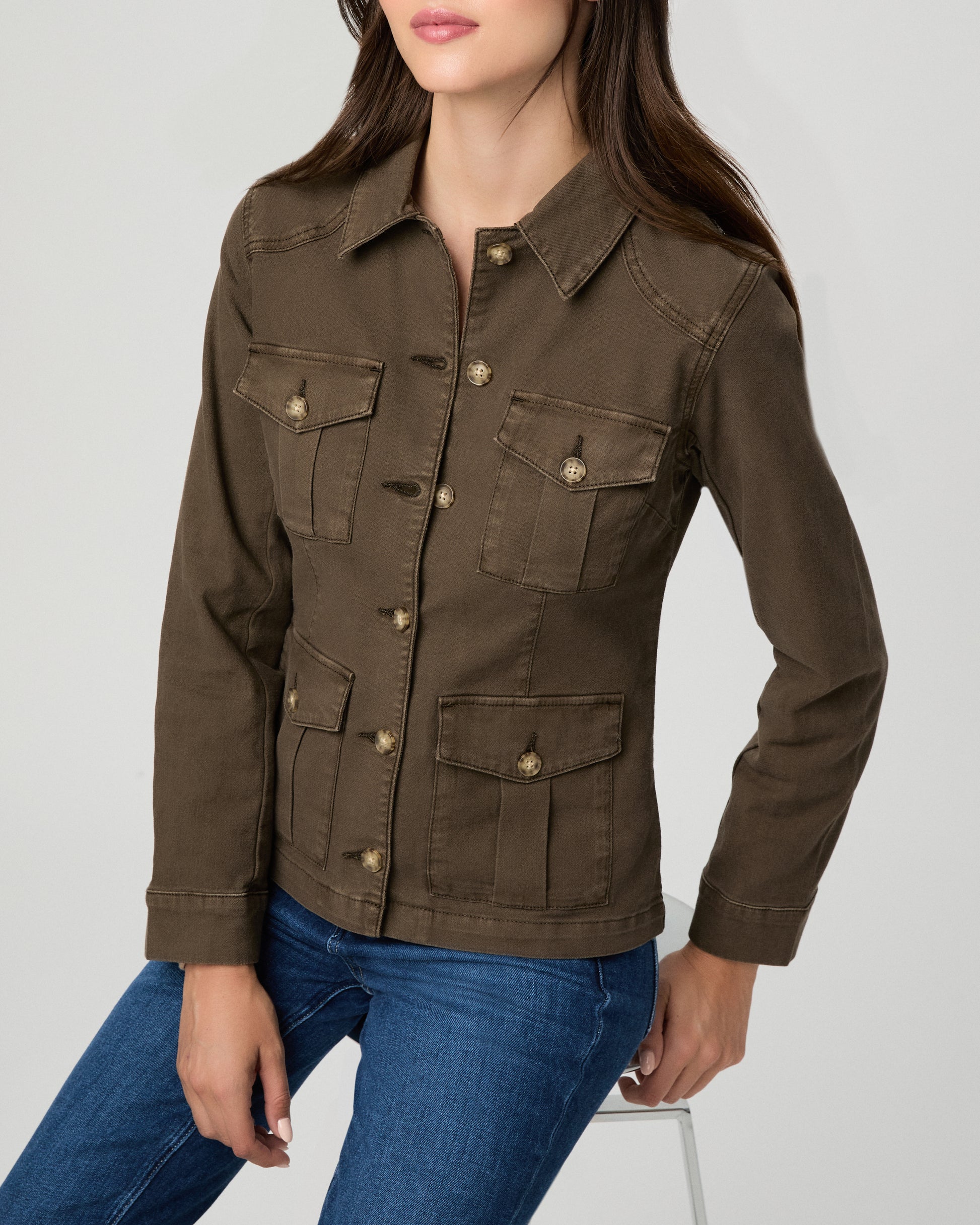 A woman modeling a dark brown utility jacket with four front flap pockets and gold-tone buttons, paired with blue jeans, against a light gray studio background.