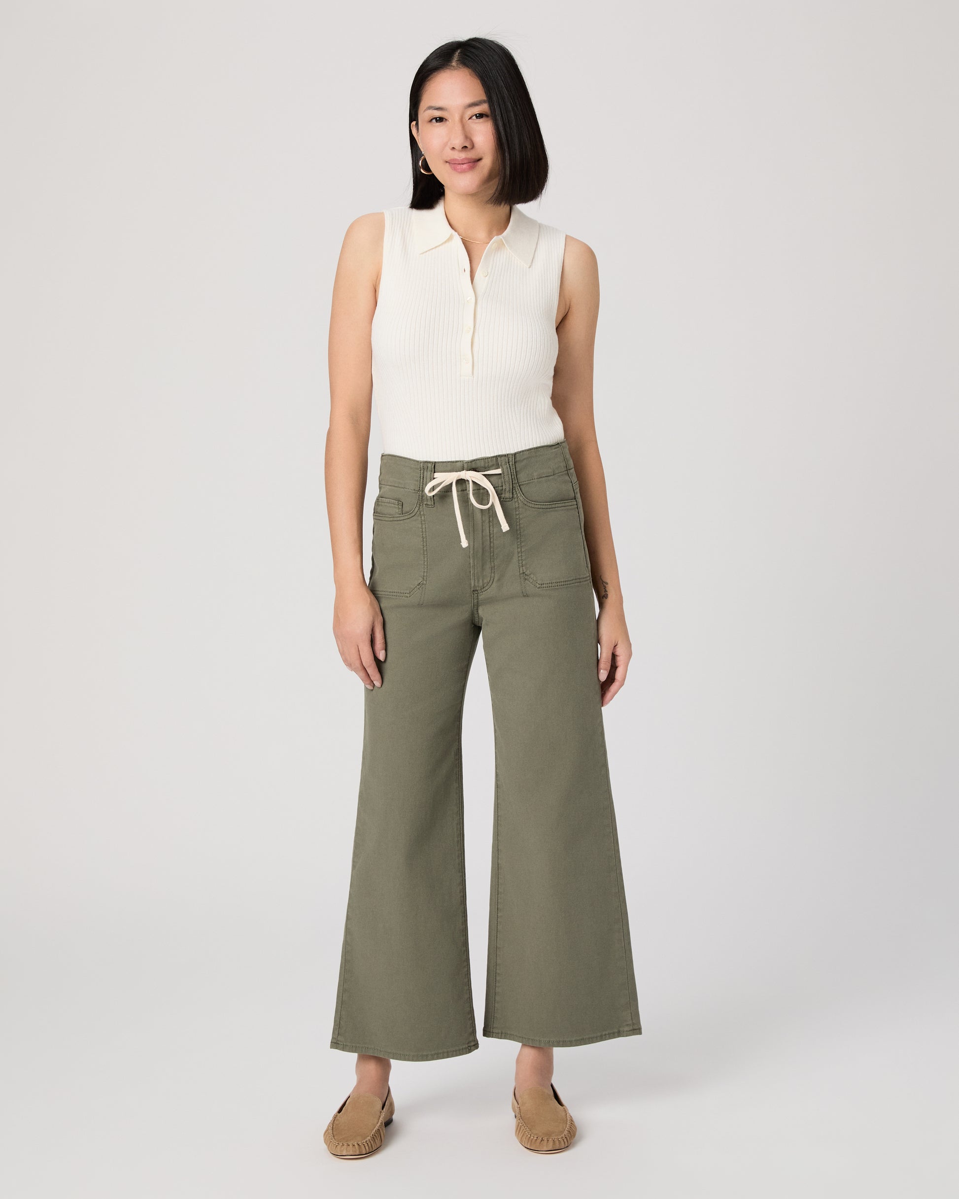 Woman wearing a white sleeveless top and green pants paired with brown flats on a white background