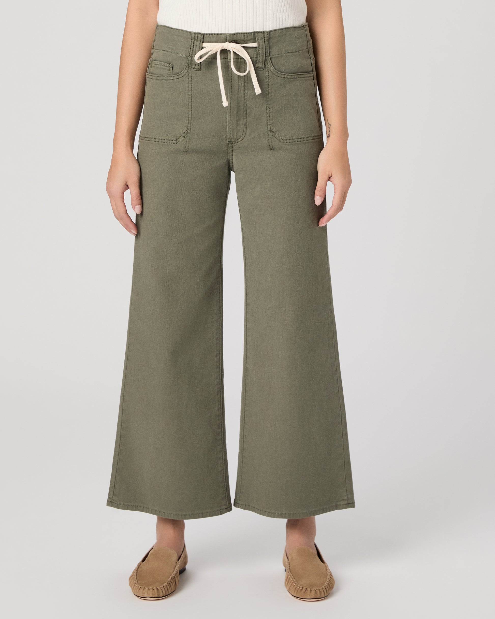 Woman wearing a white sleeveless top and green pants paired with brown flats on a white background