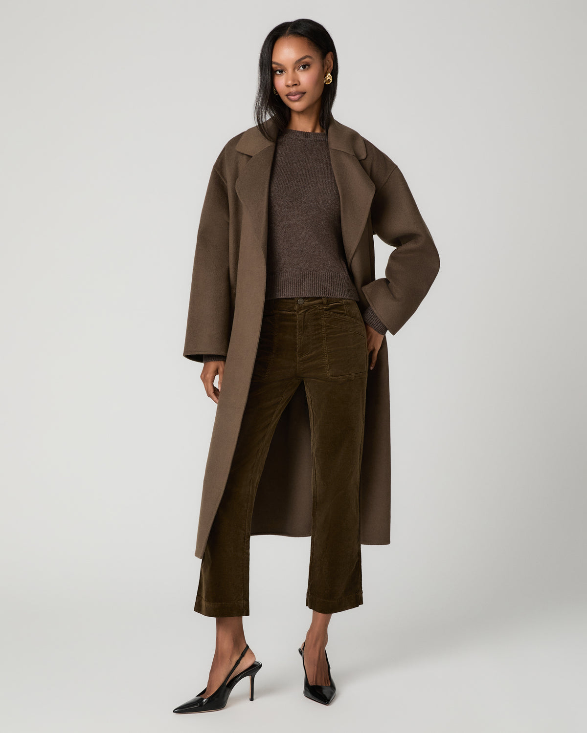 Full-length front view of a woman wearing a long olive coat over a brown sweater and fern green corduroy straight-leg ankle pants, with one hand in her pocket, paired with black pointed-toe heels.