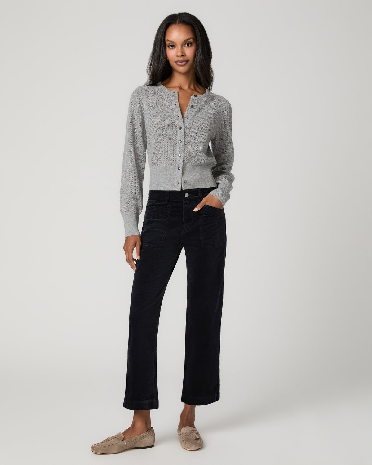 Full-length front view of a woman wearing a gray cardigan and navy corduroy straight-leg ankle pants with one hand in her pocket, paired with tan mule flats.