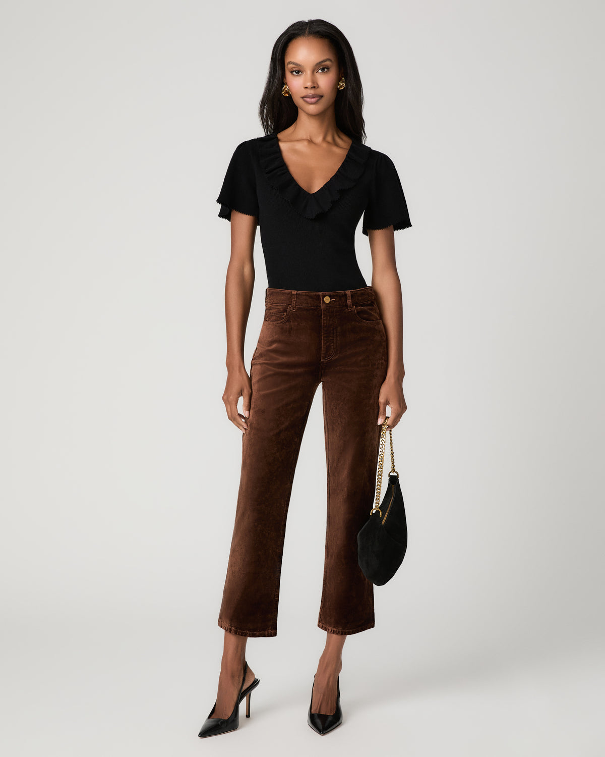 Full-length front view of a woman wearing rich brown velvet ankle-length straight jeans with a black ruffled short-sleeve top, holding a black clutch and wearing black pointed heels.