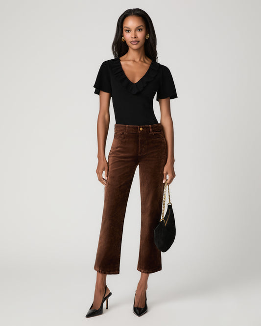 Full-length front view of a woman wearing rich brown velvet ankle-length straight jeans with a black ruffled short-sleeve top, holding a black clutch and wearing black pointed heels.