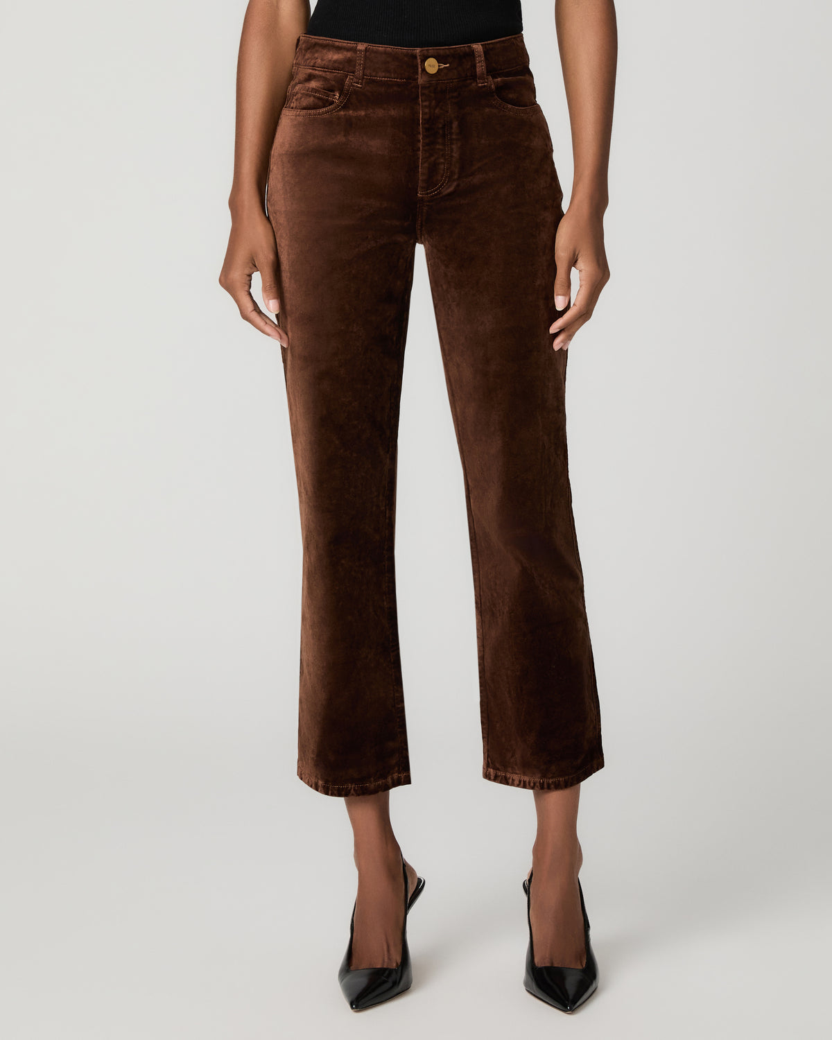Close-up front view of a woman's legs in rich brown velvet straight-leg ankle jeans, paired with black pointed pumps.