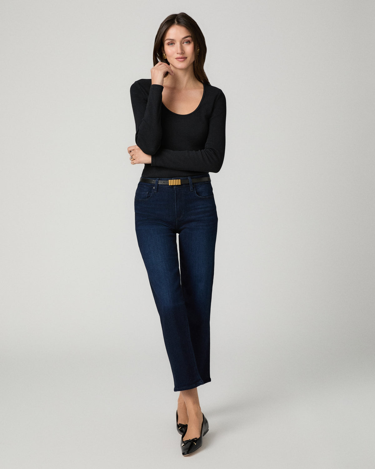 A woman modeling a black long-sleeve scoop-neck top and dark blue straight-leg jeans with a gold-tone belt, standing against a light gray studio background.
