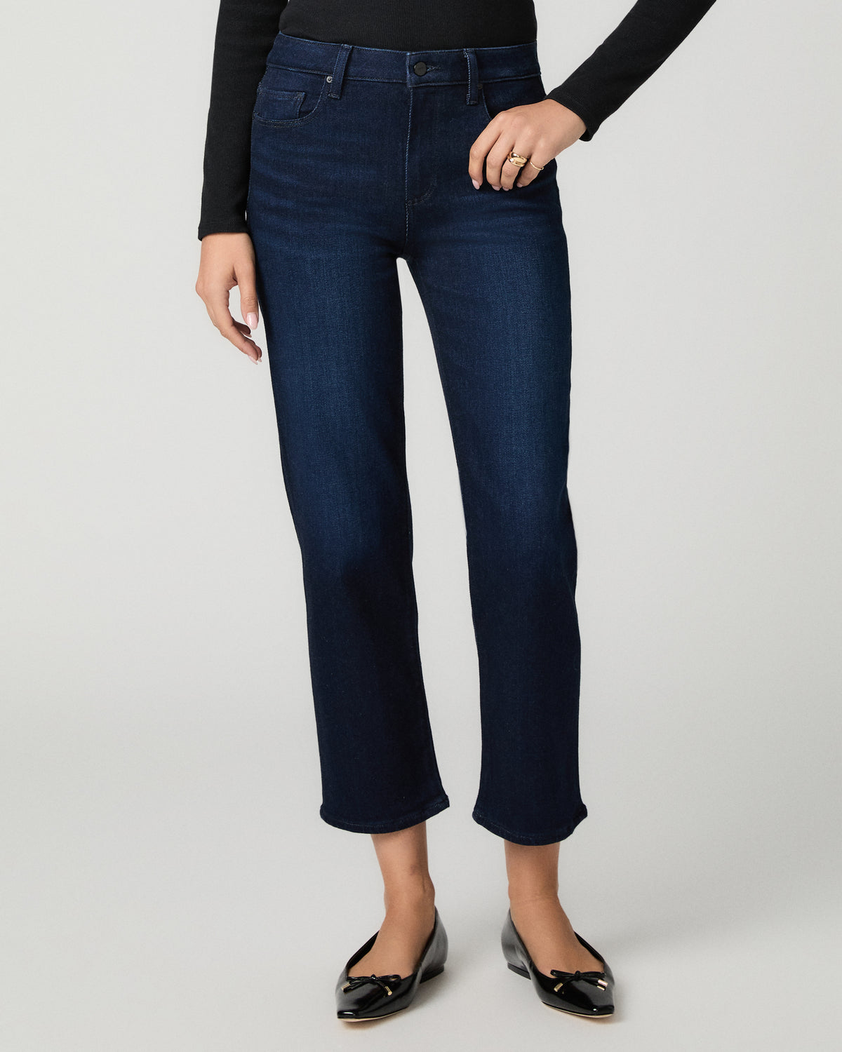 A model wearing dark blue cropped jeans with a straight leg, paired with a black long-sleeve top and black patent leather flats, shown against a light gray studio background.