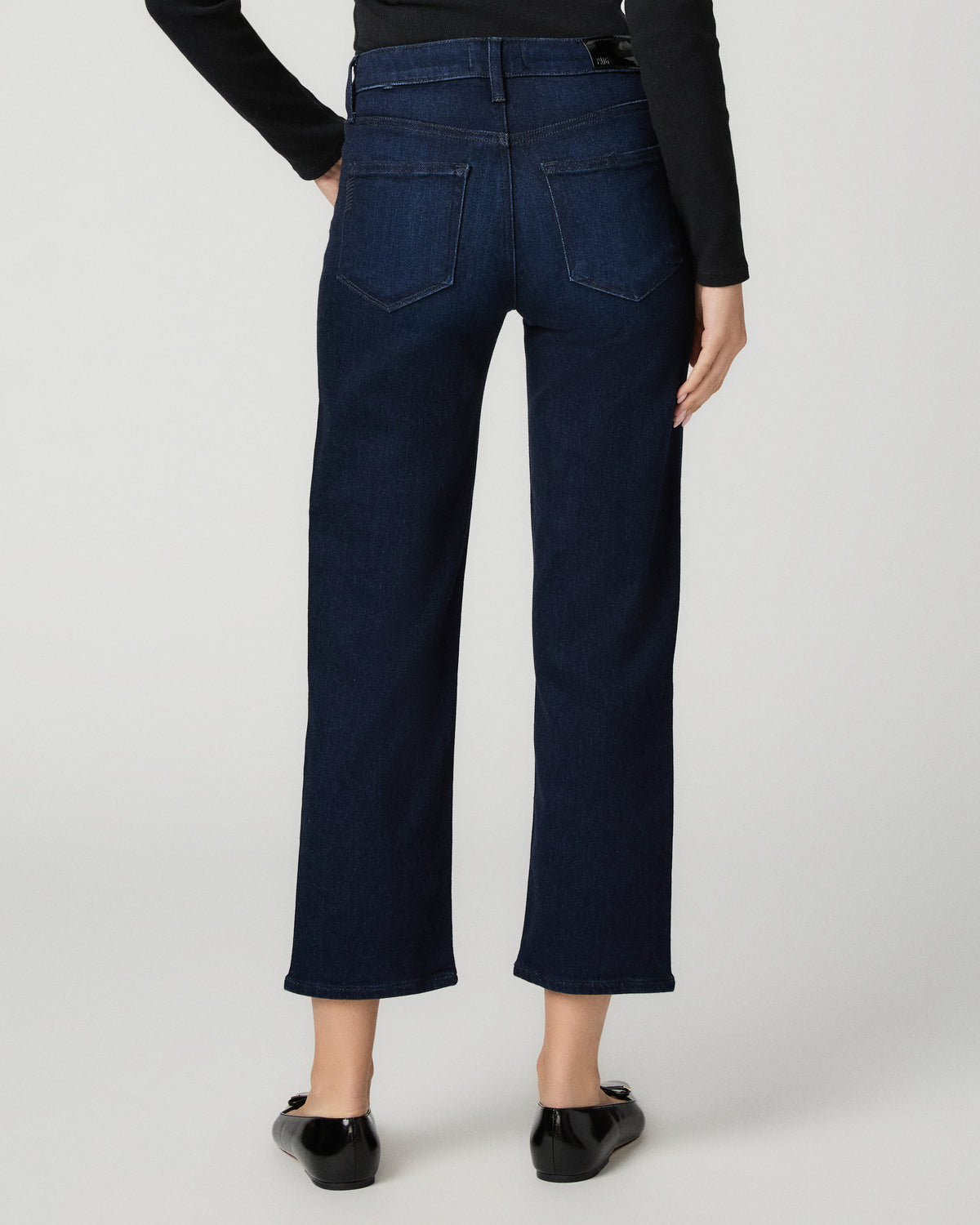 Rear view of a model wearing dark blue cropped wide-leg jeans with a black long-sleeve top and black flats, set against a light gray studio background.