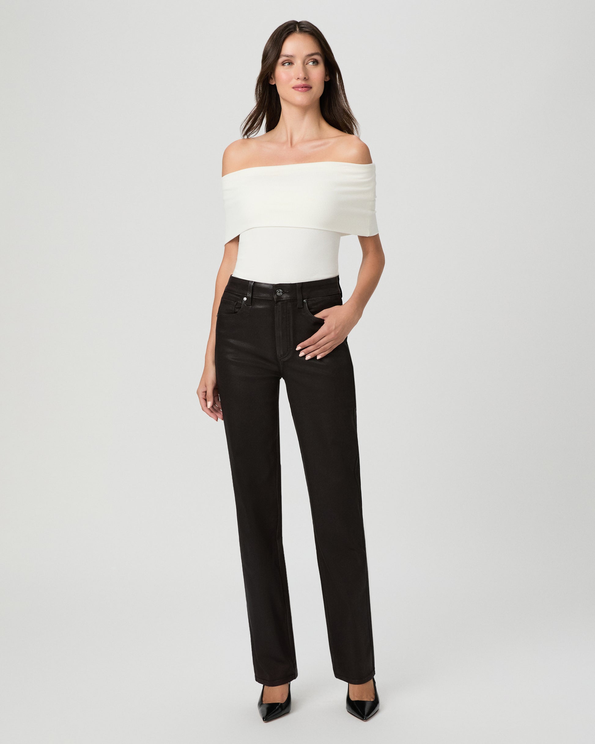 A woman modeling an off-the-shoulder white top and black straight-leg jeans against a light gray studio background.