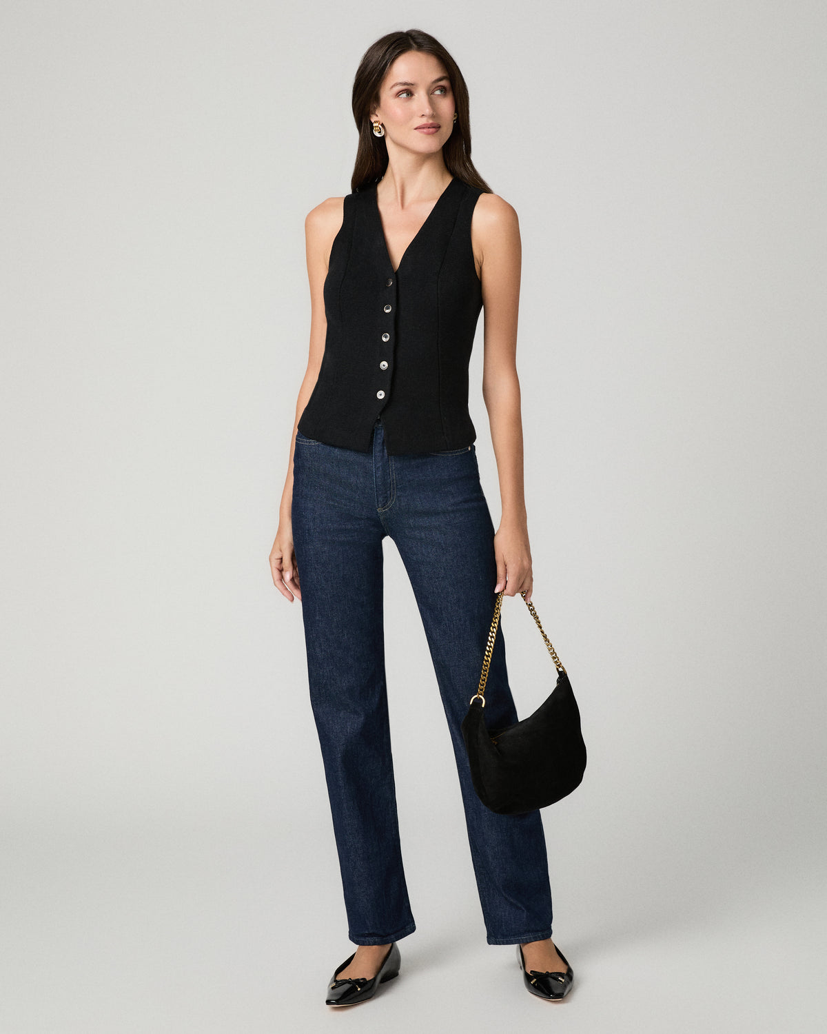 A woman modeling a sleeveless black button-front vest over a black top, paired with dark blue jeans, black flats, and holding a black crescent shoulder bag with a gold chain strap, against a light gray studio background.