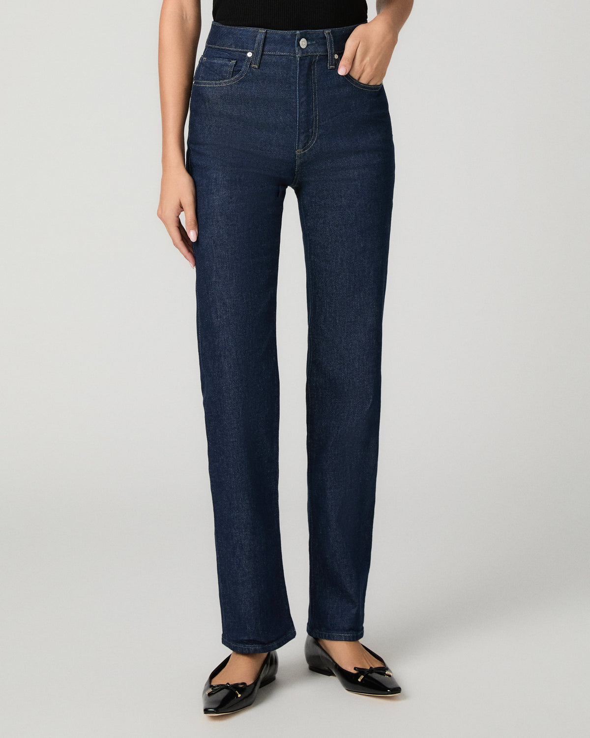 A model wearing a pair of dark blue straight-leg jeans, paired with a black top and black patent leather flats, photographed against a neutral light gray studio background.