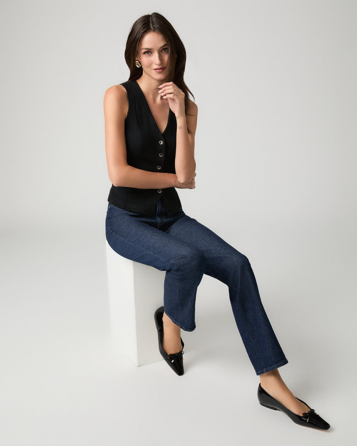 A woman sits on a white block in a studio setting, wearing a black sleeveless button-front top, dark blue straight-leg jeans, and black flats.