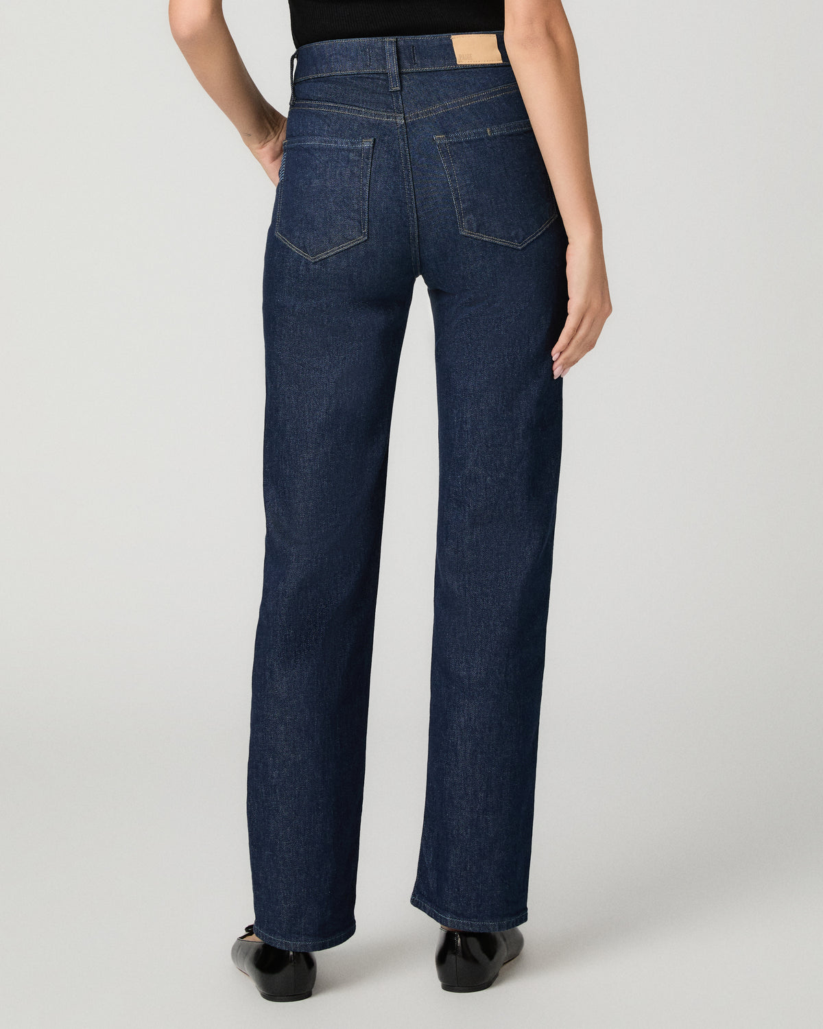 Rear view of a model wearing dark blue straight-leg denim jeans with a black top and black shoes, photographed against a light gray studio background.