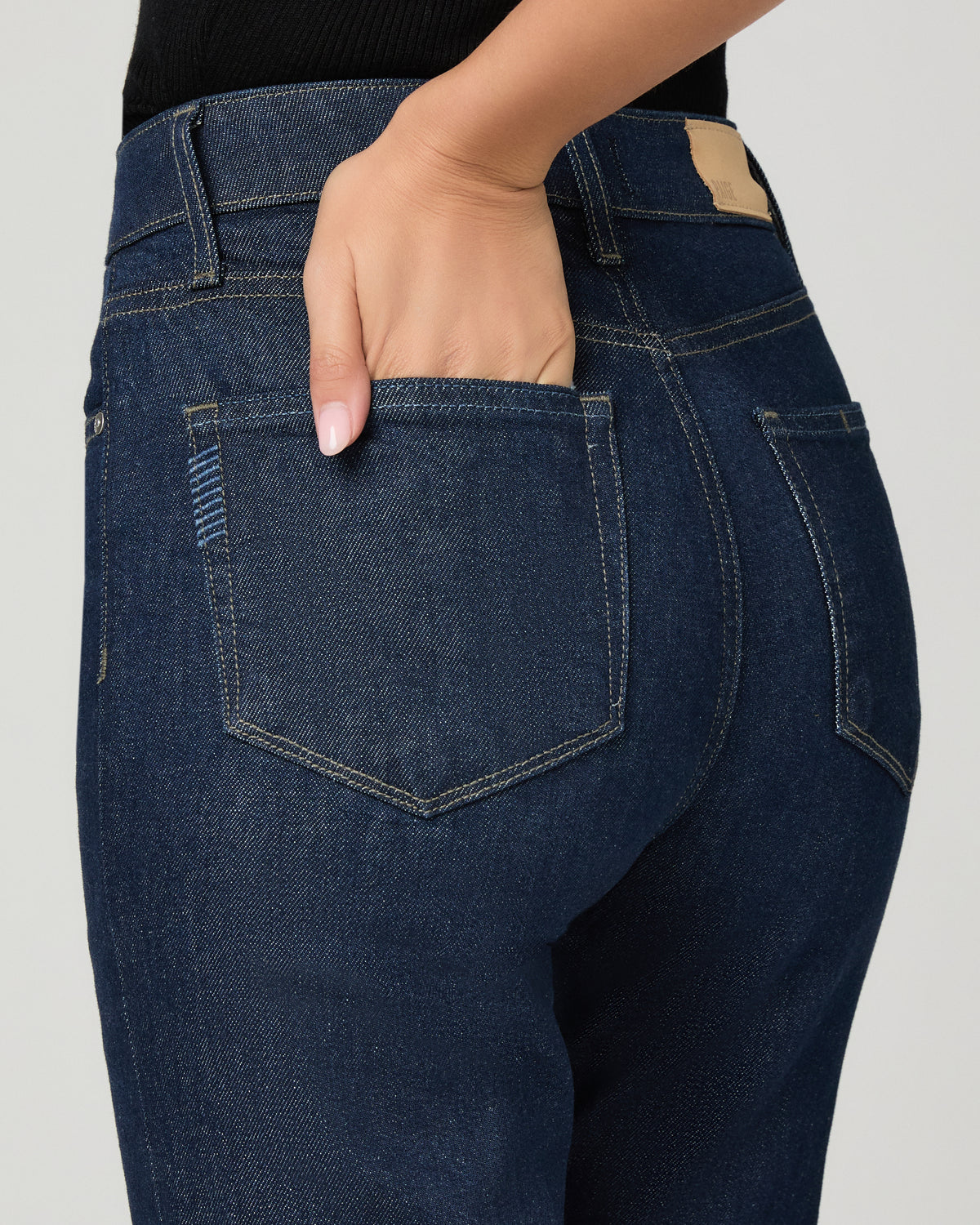 Close-up back view of a person wearing dark indigo denim jeans, with one hand tucked into the back pocket. The photo shows the waistband, belt loop, back pocket construction, and a small tan brand patch near the waistband against a light gray studio background.