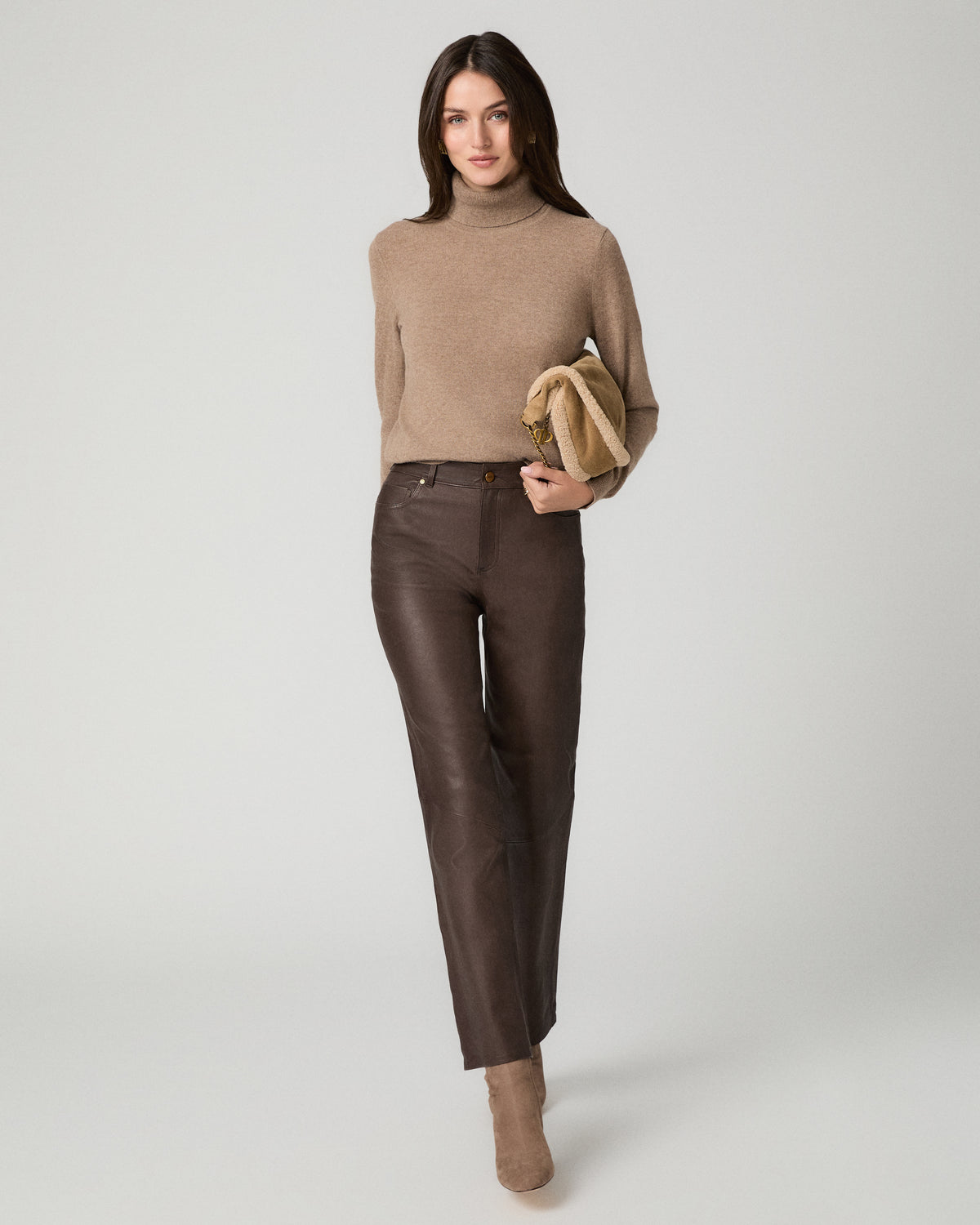A woman modeling a beige knit turtleneck sweater, dark brown leather trousers, and beige ankle boots, holding a small beige clutch against a light gray studio background.