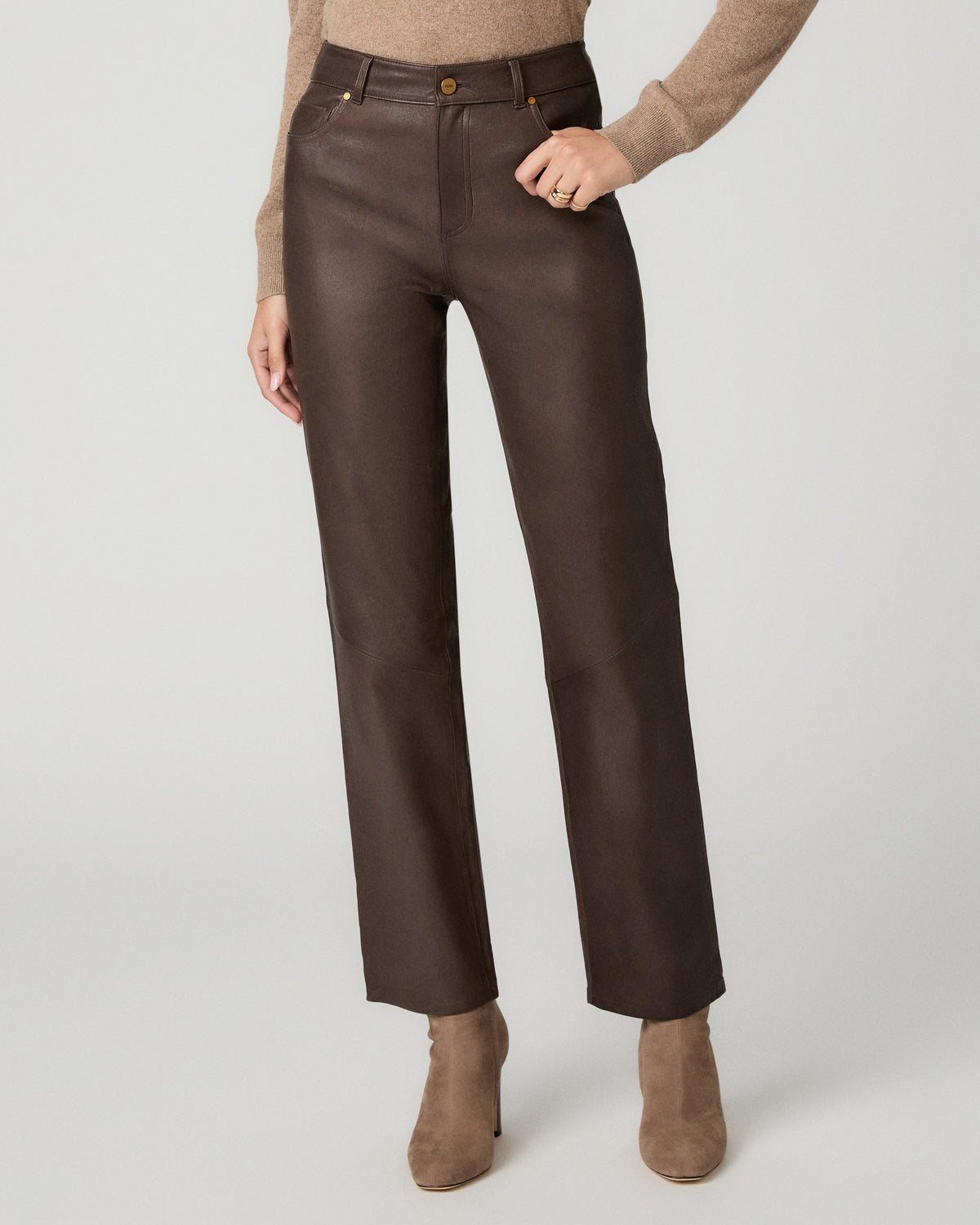 A model wearing dark brown faux-leather straight-leg pants, styled with a beige knit sweater and taupe suede ankle boots against a light gray studio background.