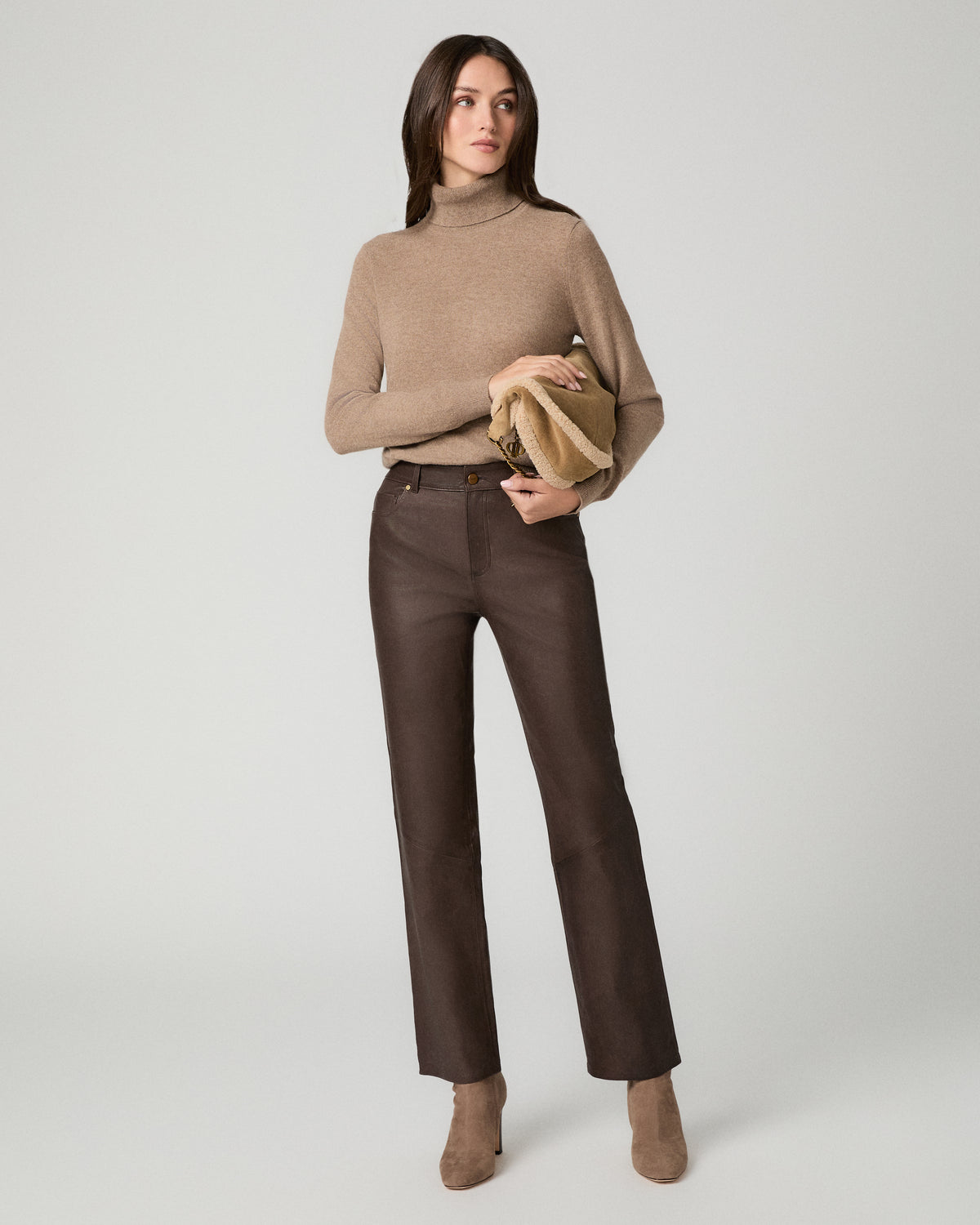 A woman modeling a beige mock-neck knit sweater and dark brown straight-leg trousers, holding a tan clutch against a light gray studio background.