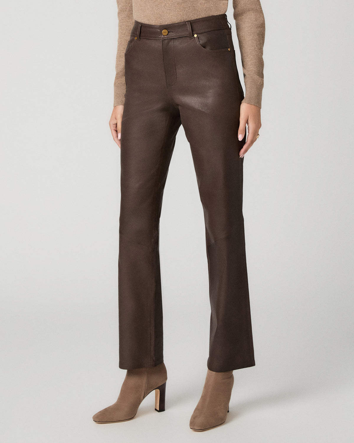A woman modeling a beige knit turtleneck sweater, dark brown leather trousers, and beige ankle boots, holding a small beige clutch against a light gray studio background.