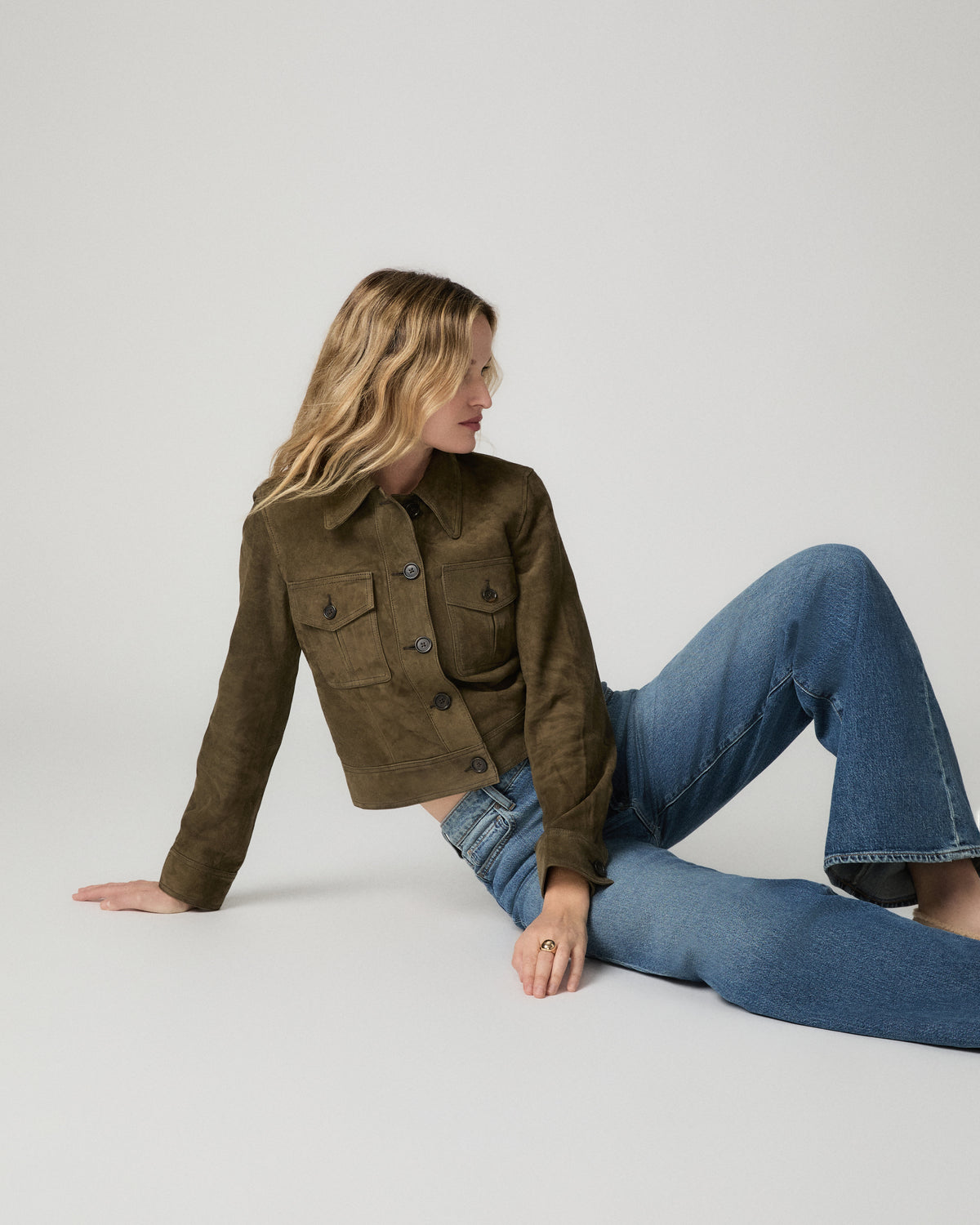 Woman seated on the ground wearing the rich olive suede Feyd Jacket with blue denim jeans.