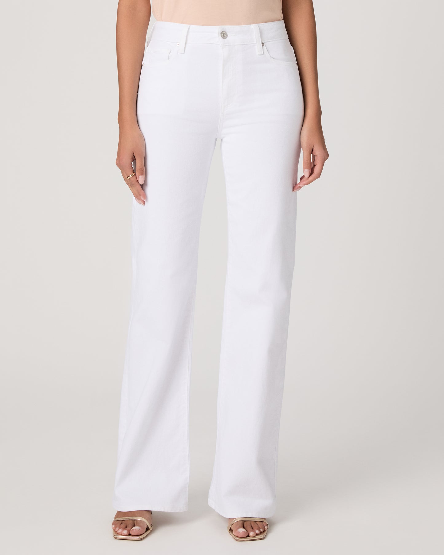 Woman wearing a light pink sleeveless top and white pants paired with gold sandals on a plain background