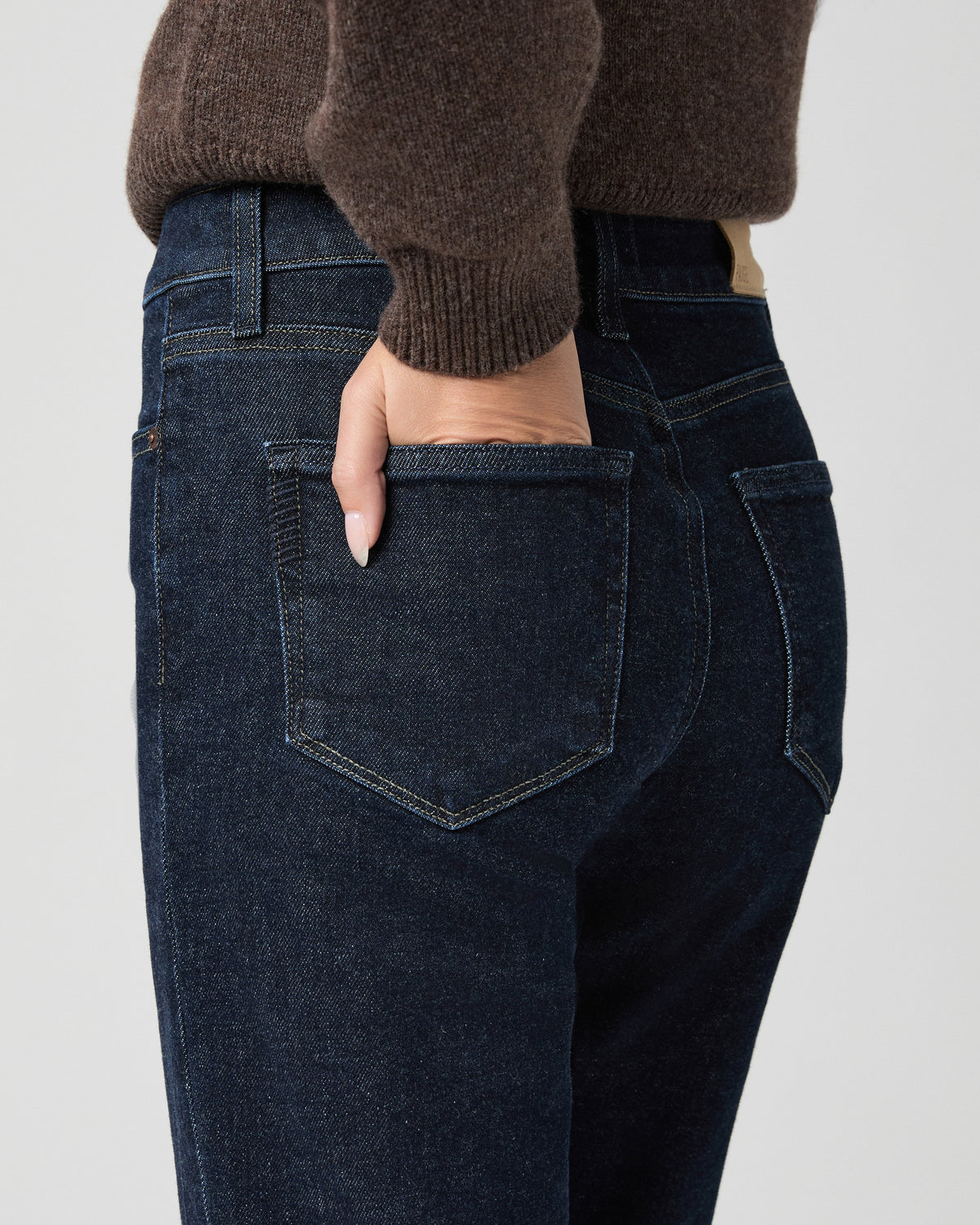 Close-up of the back pocket of the Stevie Ankle Straight Jean in Moderne dark wash; model has one hand in the pocket and wears a brown sweater