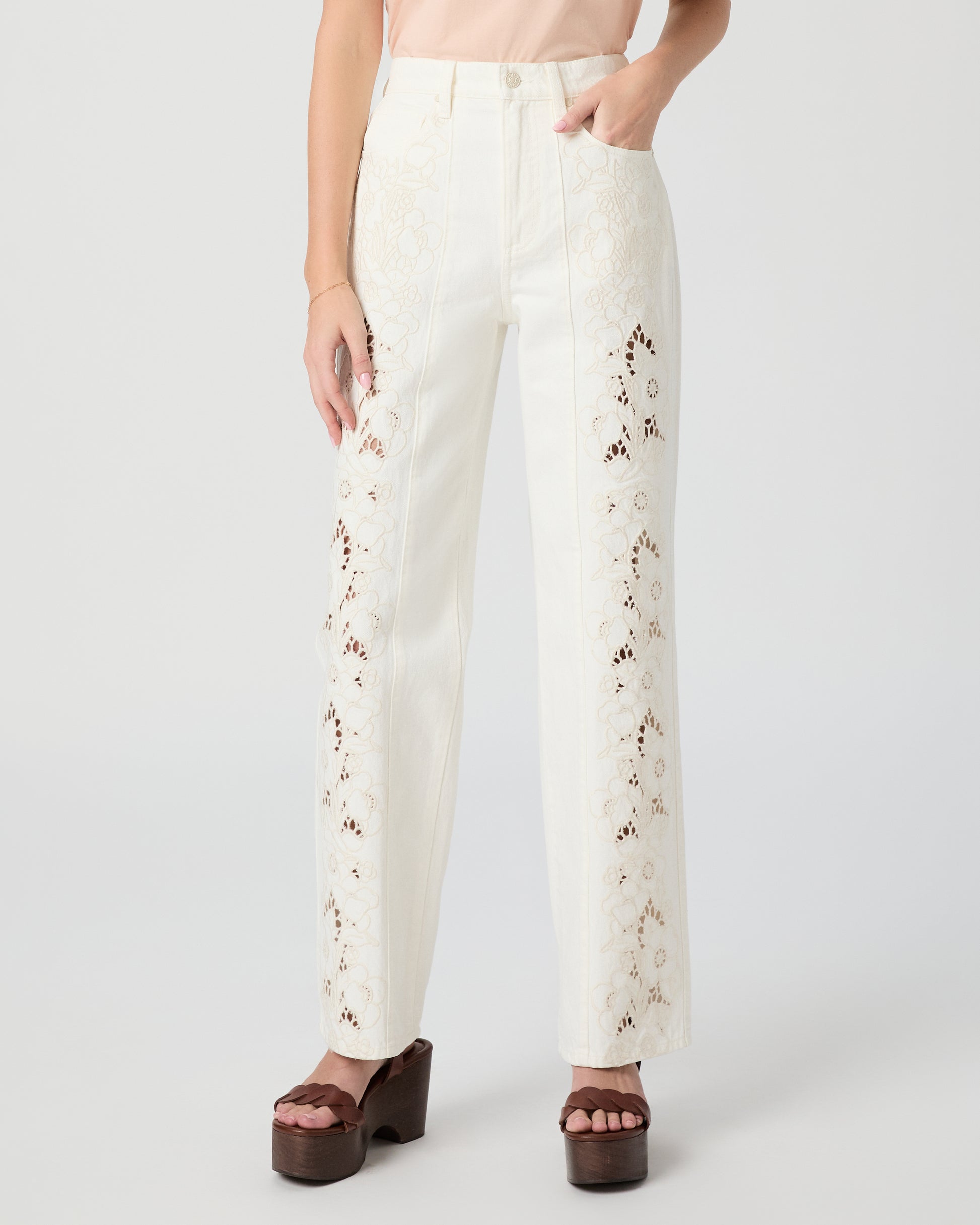 Woman wearing a pink top and white pants paired with brown heels on a plain background