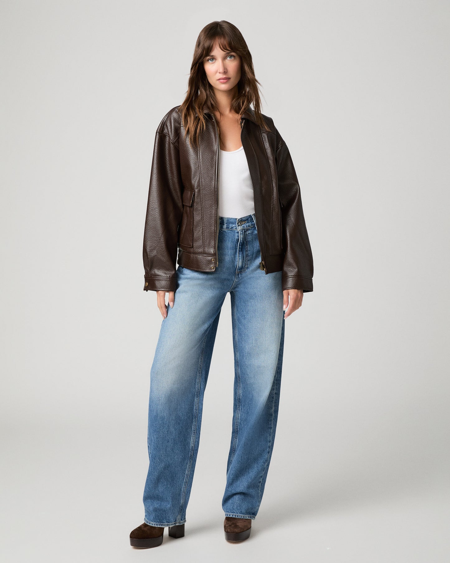 Woman in dark brown jacket and a white top with light blue jeans and a white background