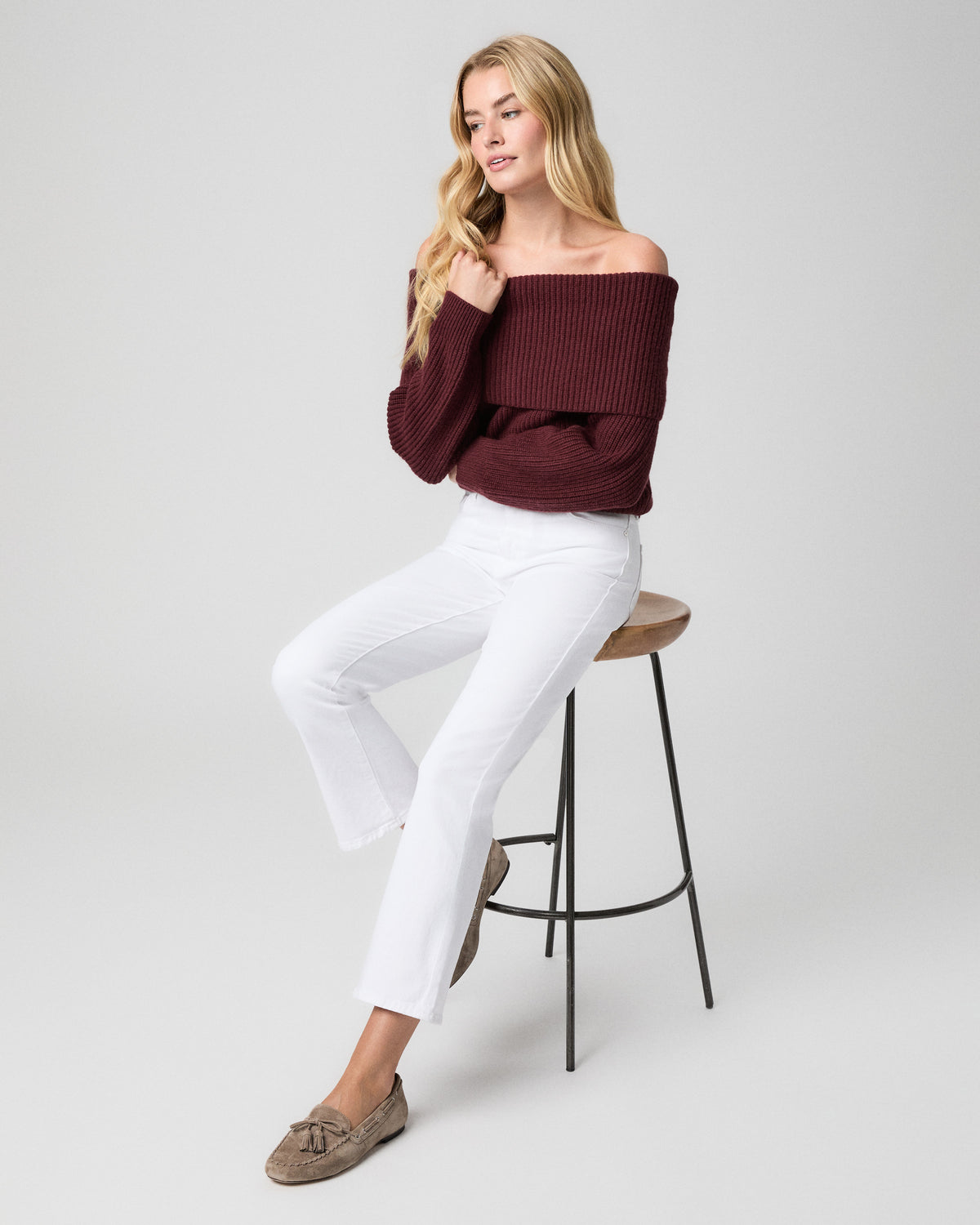 Model seated on a wooden stool, one hand touching her hair, wearing a burgundy off-the-shoulder sweater, Mason Crop Bootcut Jeans in Radiant White, and taupe loafers