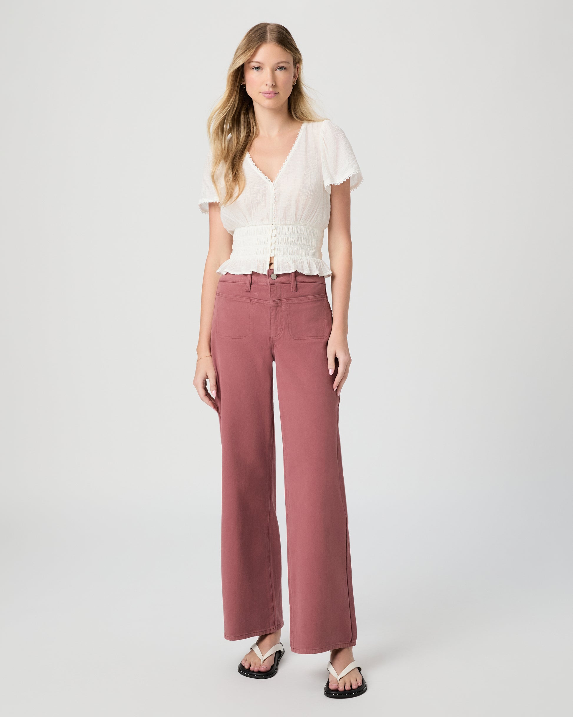 Woman wearing a white blouse and pink jeans paired with white sandals on a gray background