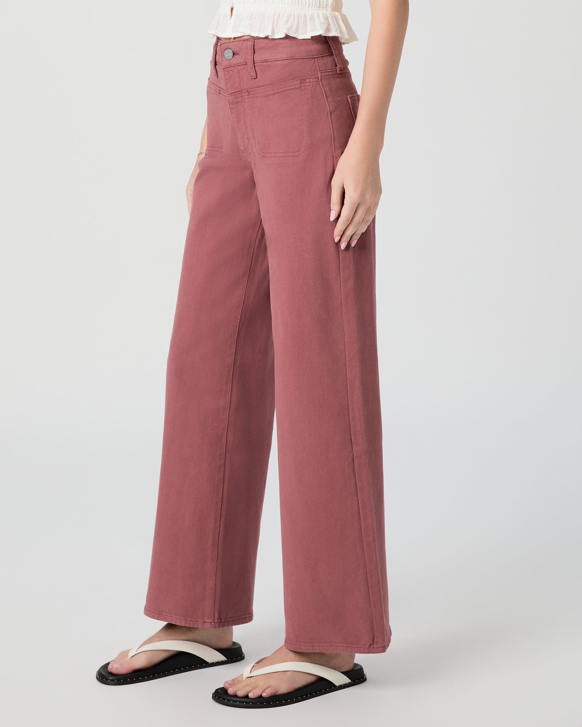Woman wearing a white blouse and pink jeans paired with white sandals on a gray background