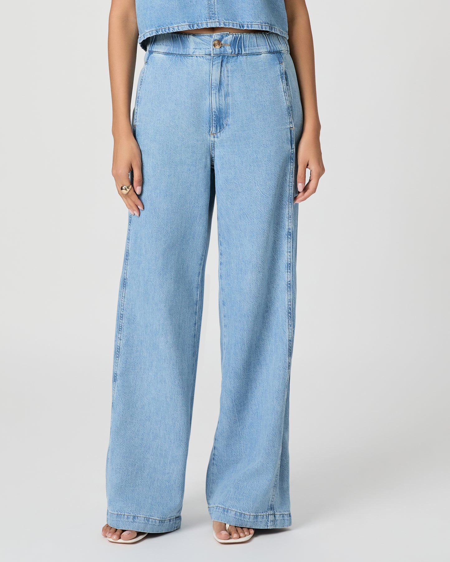 Woman wearing a light blue denim top and denim pants paired with white sandals on a white background