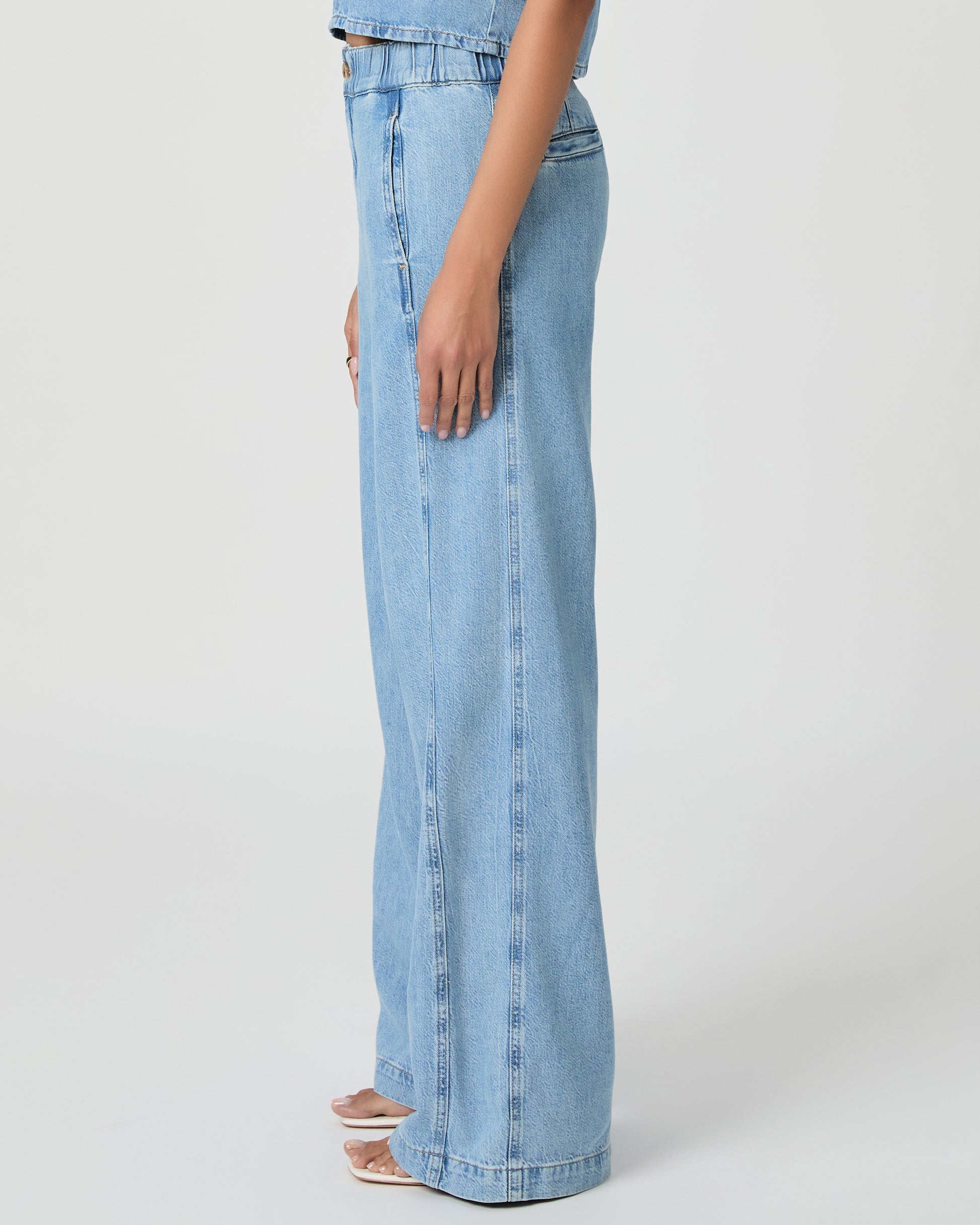 Woman wearing a light blue denim top and denim pants paired with white sandals on a white background