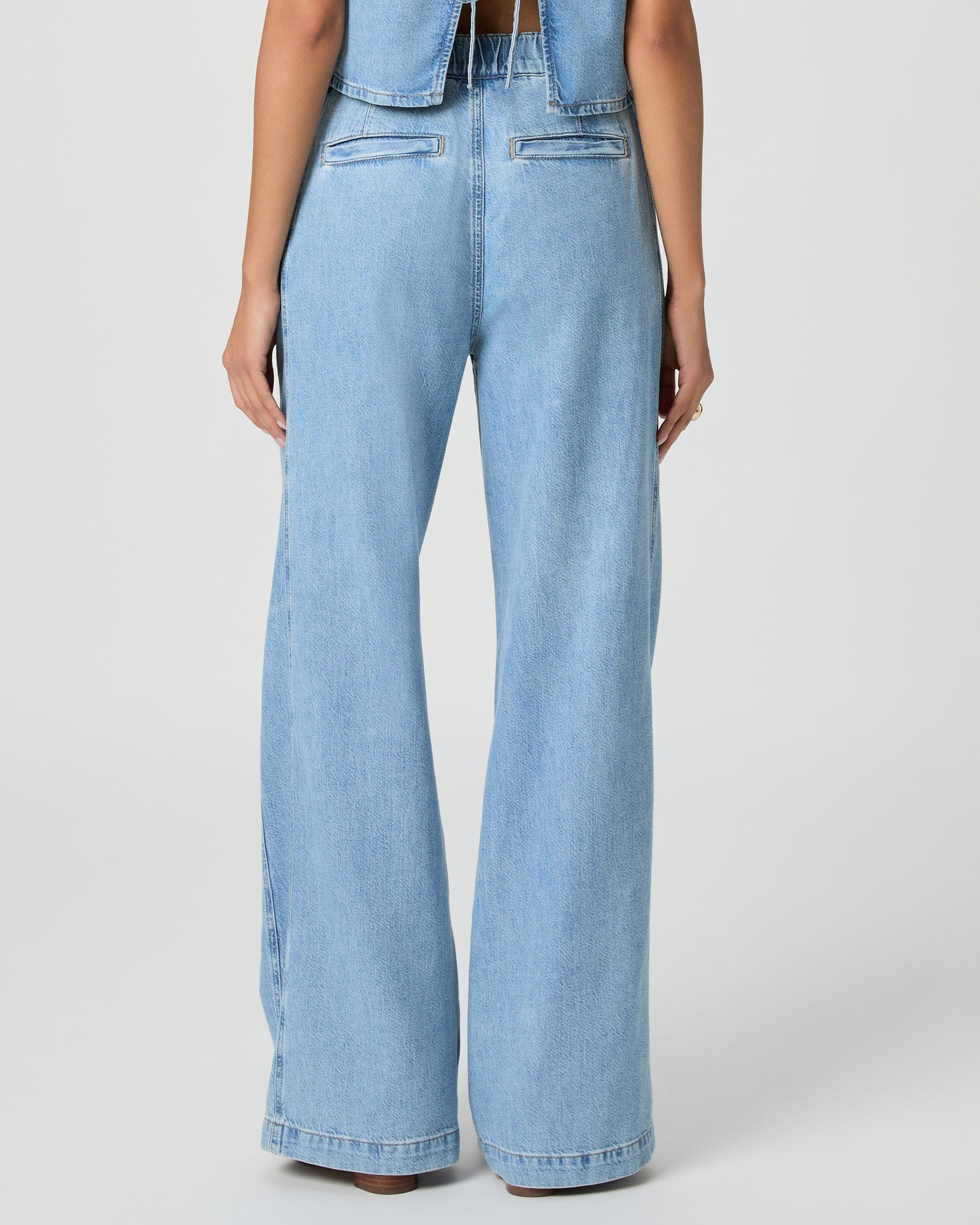 Woman wearing a light blue denim top and denim pants paired with white sandals on a white background