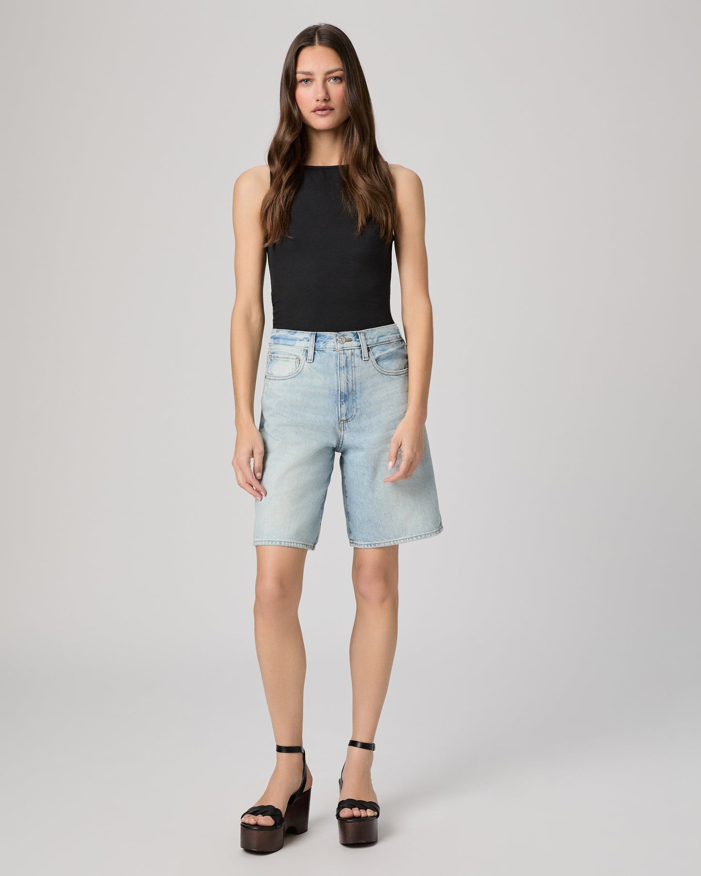 Woman wearing a black sleeveless top and light blue denim shorts paired with black heels on a gray background