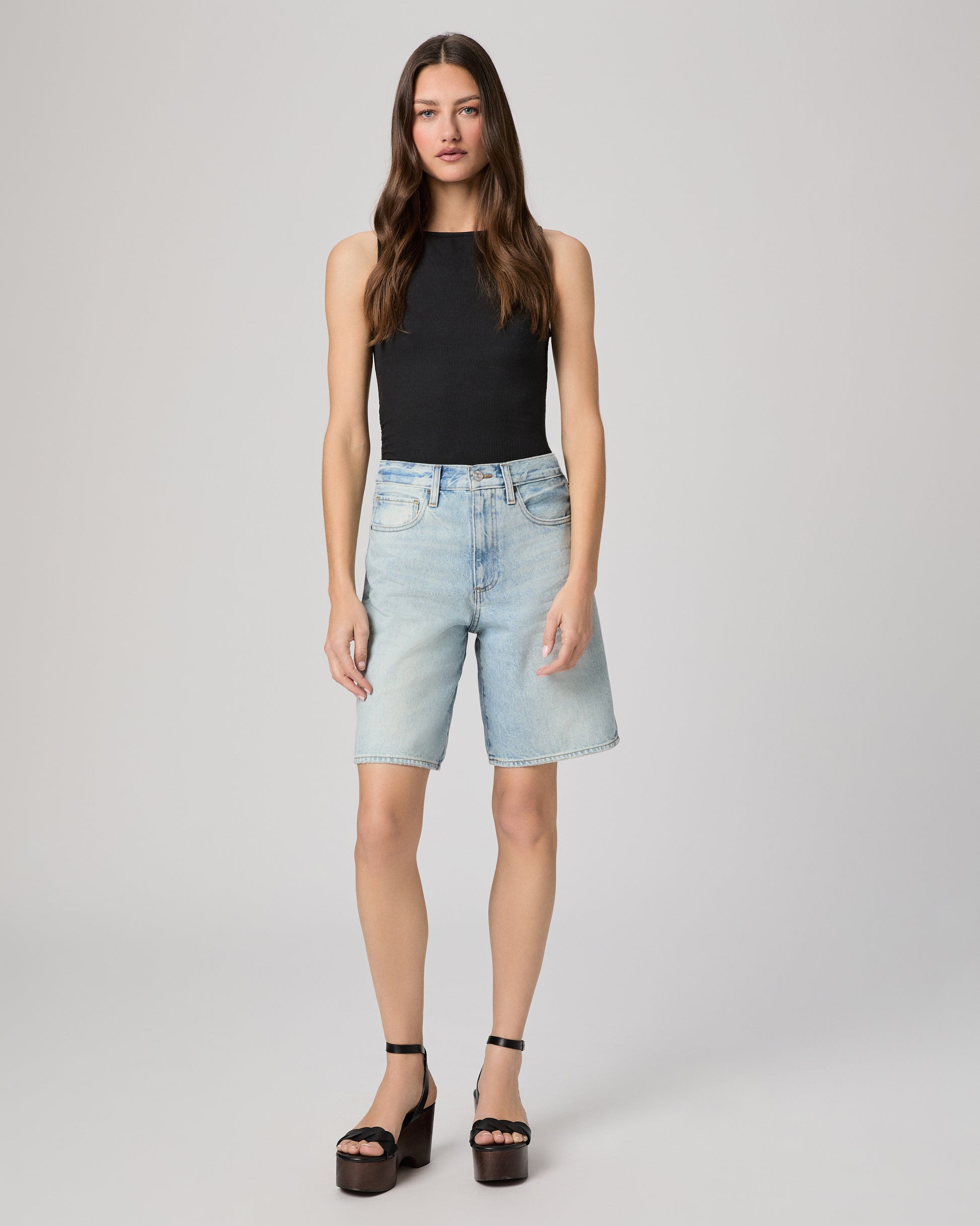 Woman wearing a black sleeveless top and light blue denim shorts paired with black heels on a gray background