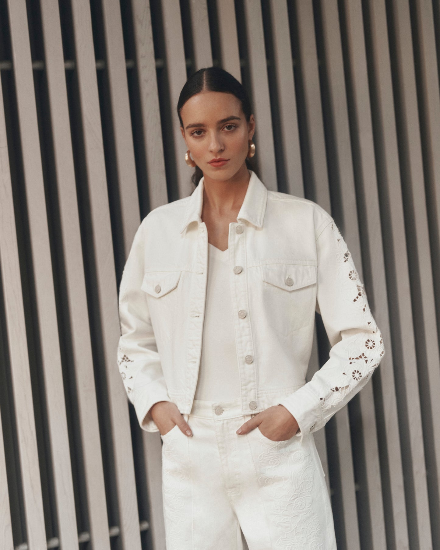Woman wearing a white denim jacket and jeans with floral embroidery against a striped wall.