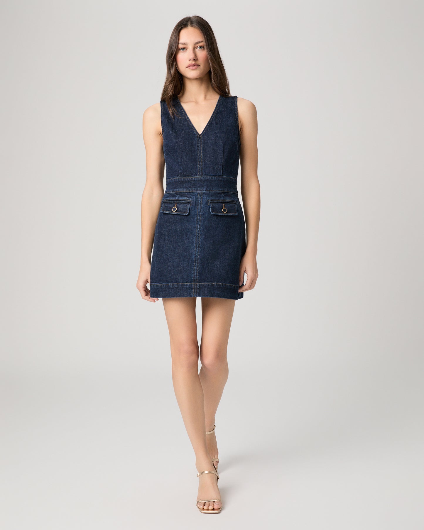 Woman wearing a mini denim dress paired with gold heels on a white background