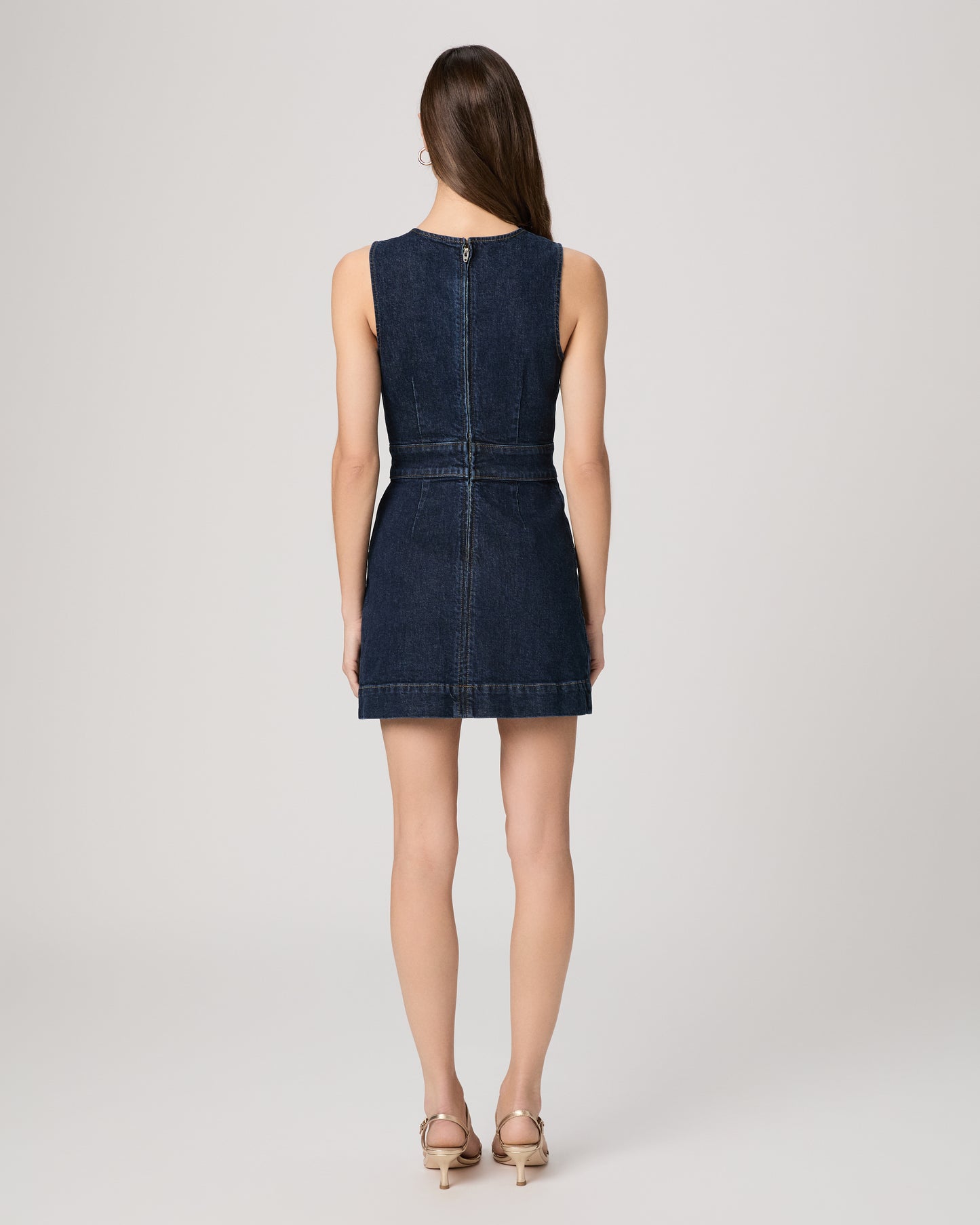Woman wearing a mini denim dress paired with gold heels on a white background