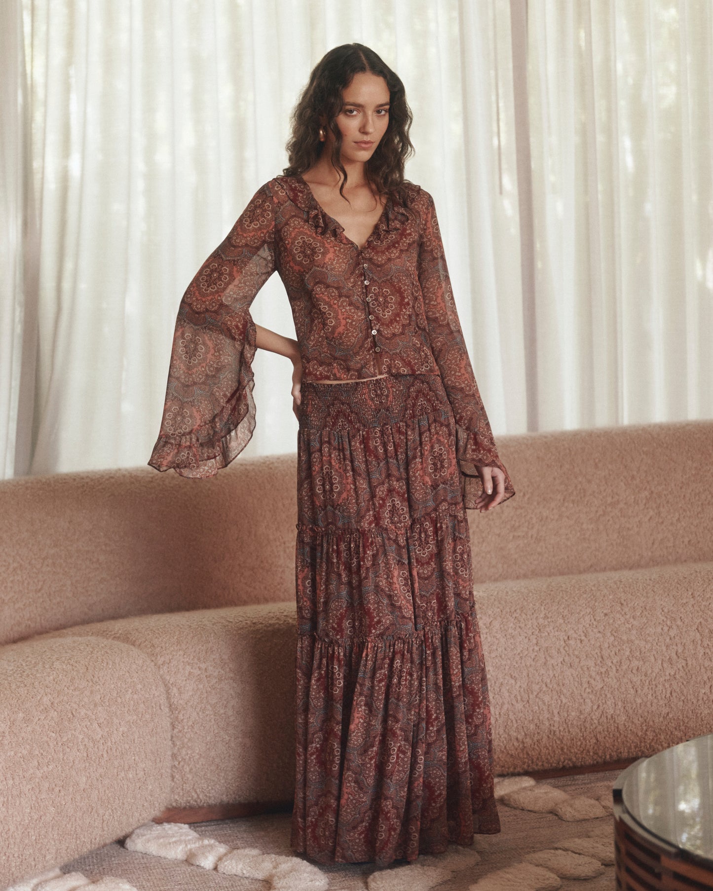 Woman wearing a long, patterned dress standing in a room with a beige sofa and white curtains.