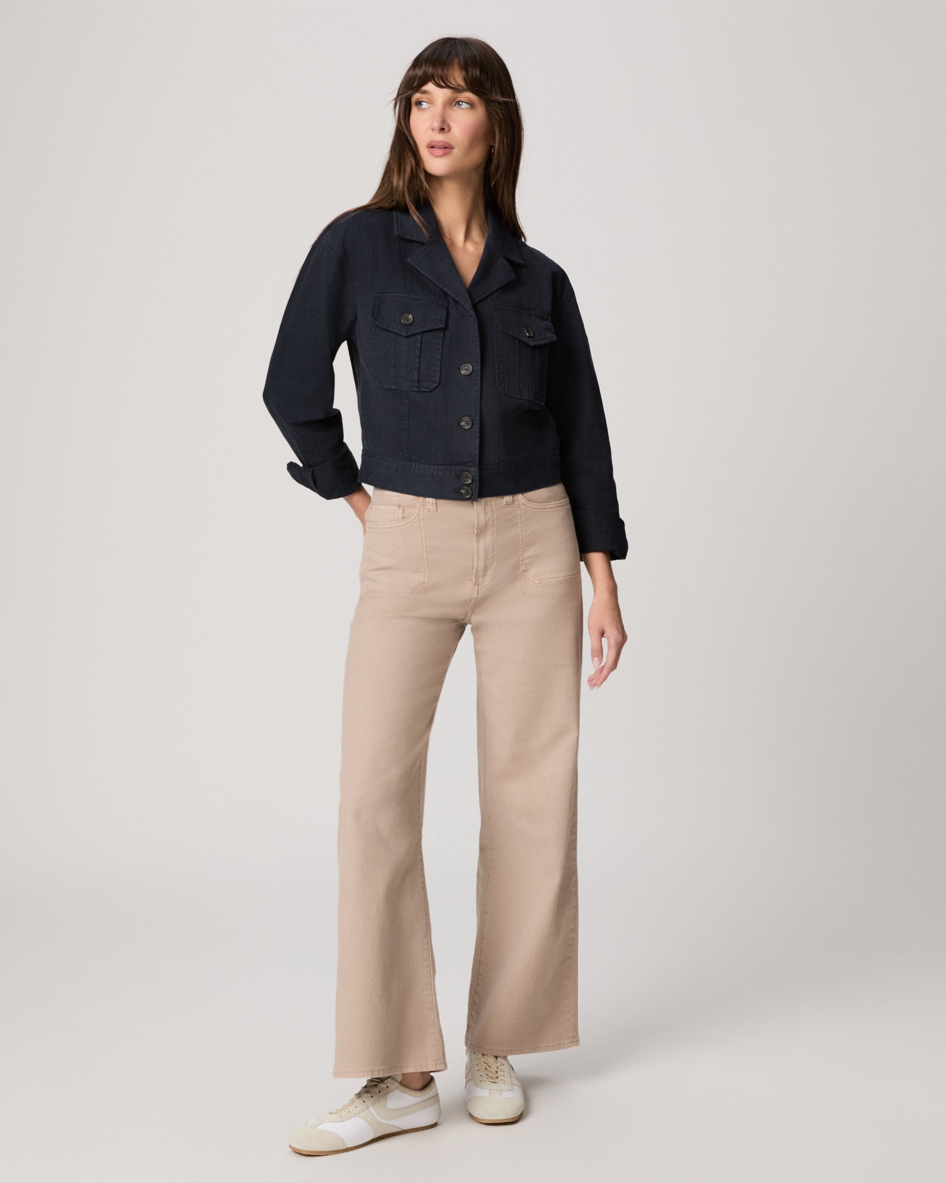 Woman wearing a navy jacket over a white top and beige pants paired with white sneakers on a light gray background